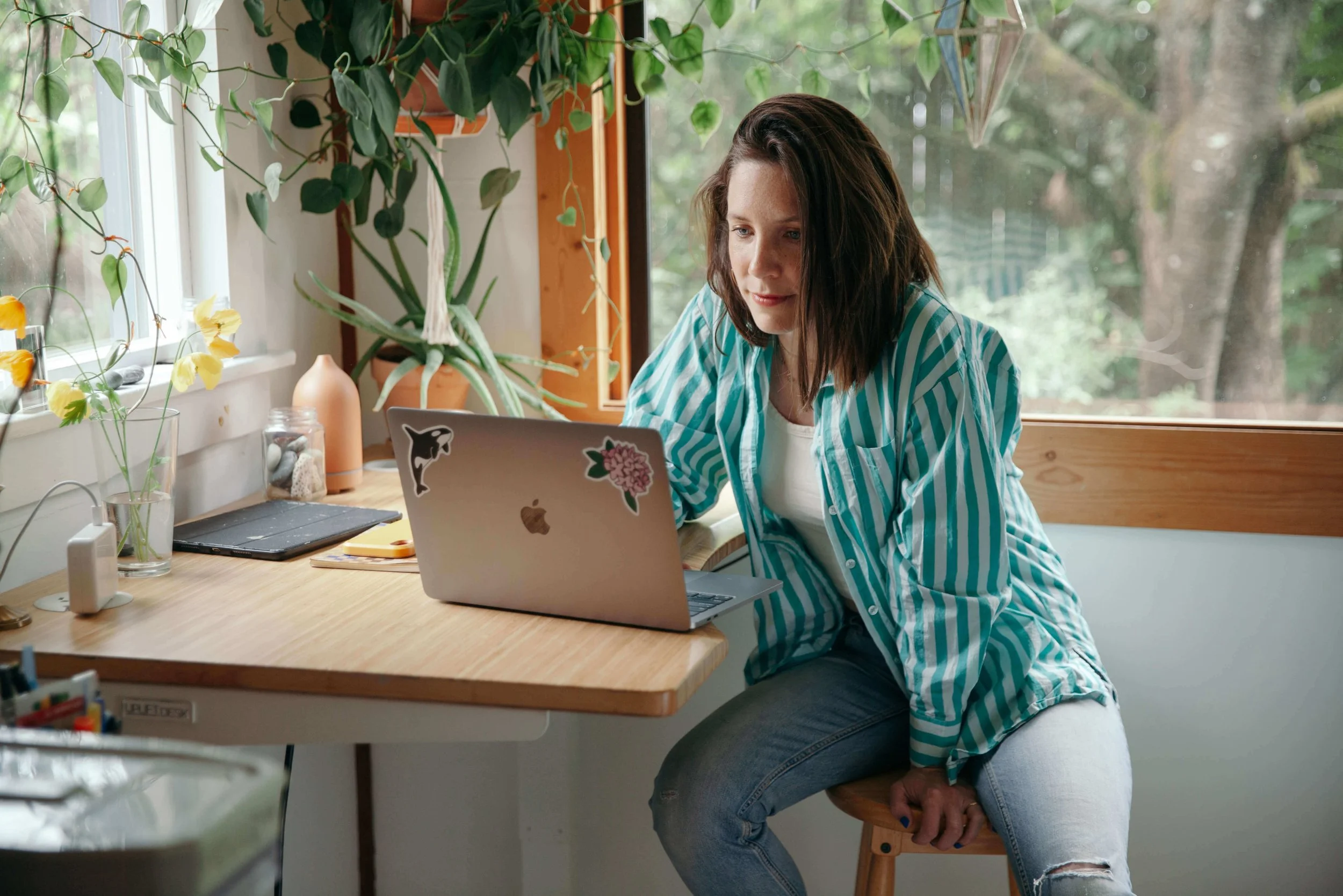 A woman with shoulder-length brown hair working on a laptop at a wooden desk in a bright room with large windows and greenery outside. The desk has various plants, a closed laptop, and decorative items.
