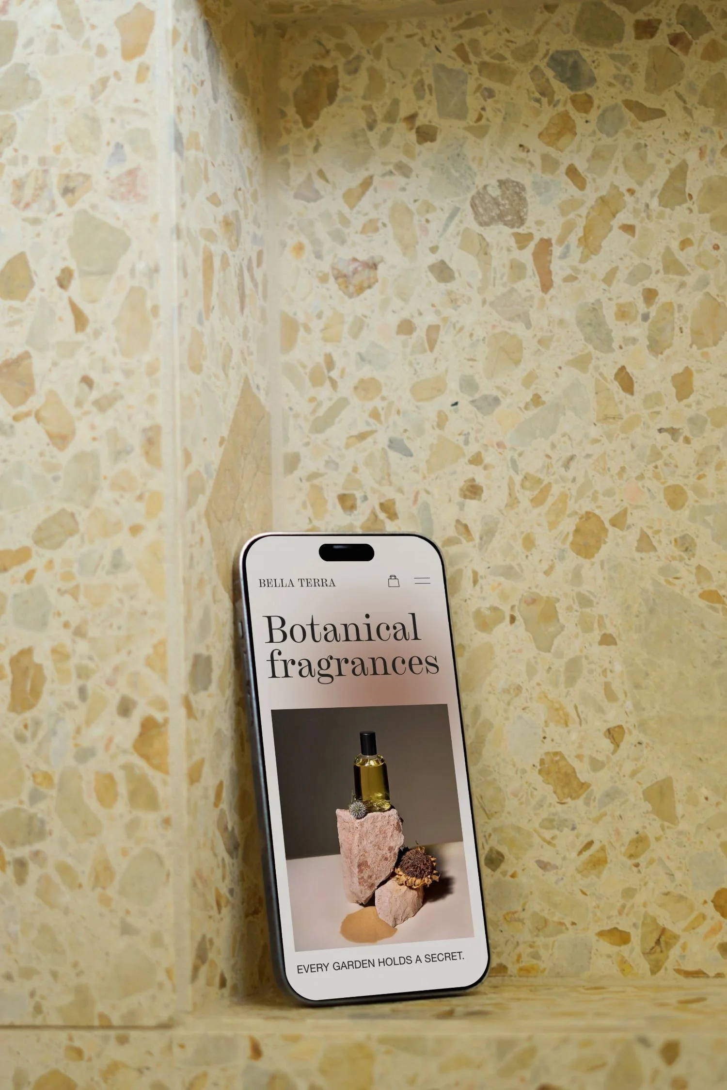 A smartphone displaying a webpage for Bella Terra Botanical fragrances, with an image of a perfume bottle placed on rocks and flowers, against a yellowish terrazzo background.