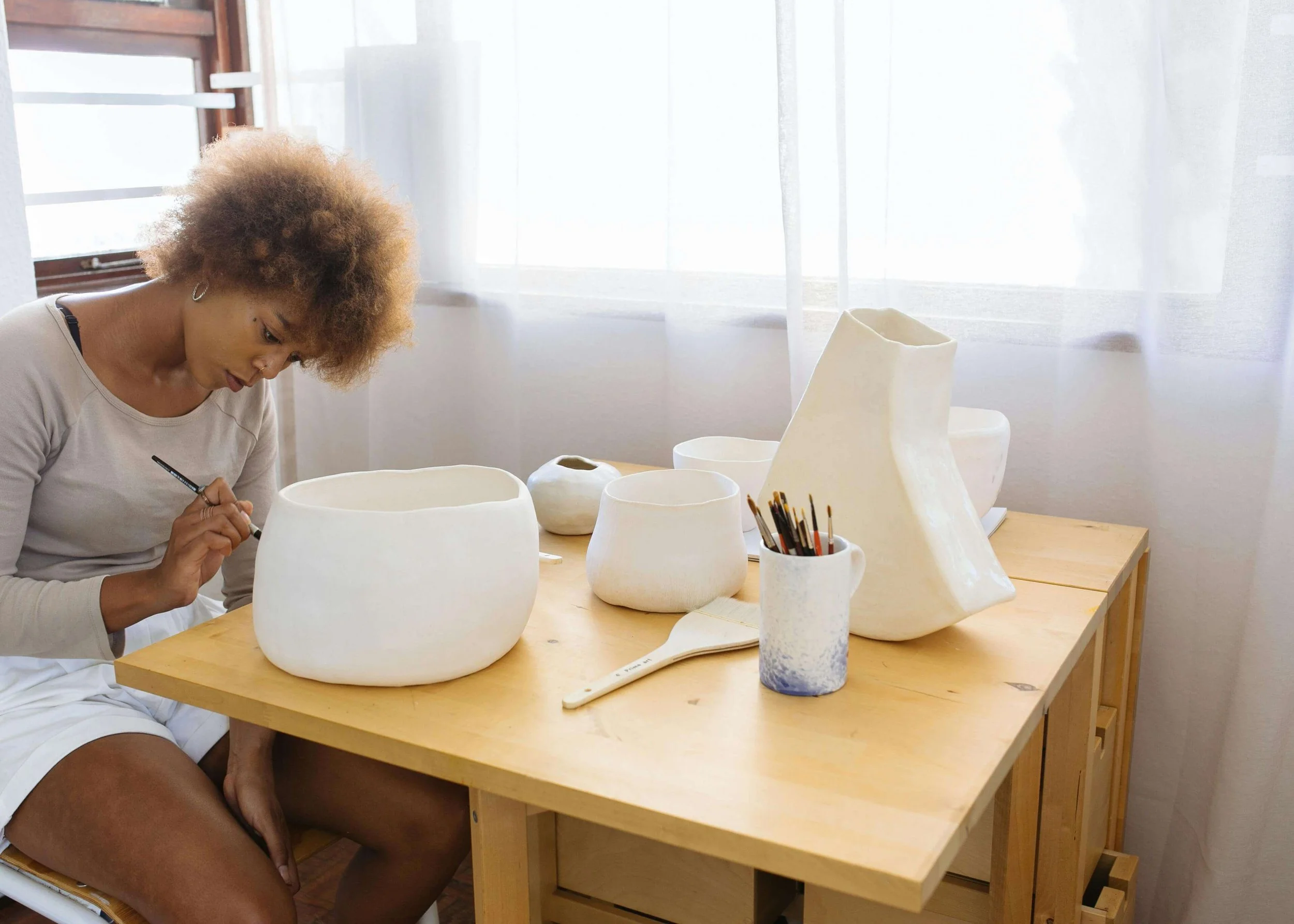 Person sitting at a table sculpting white ceramic pottery, surrounded by various clay pots and paintbrushes.