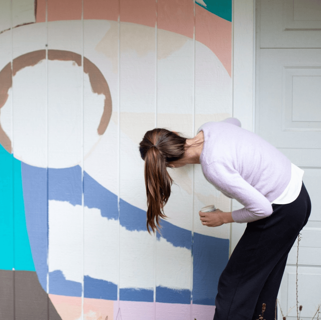 A woman with long brown hair wearing a light purple sweater and black pants is painting on a colorful mural on a vertical surface, possibly a fence or wall.