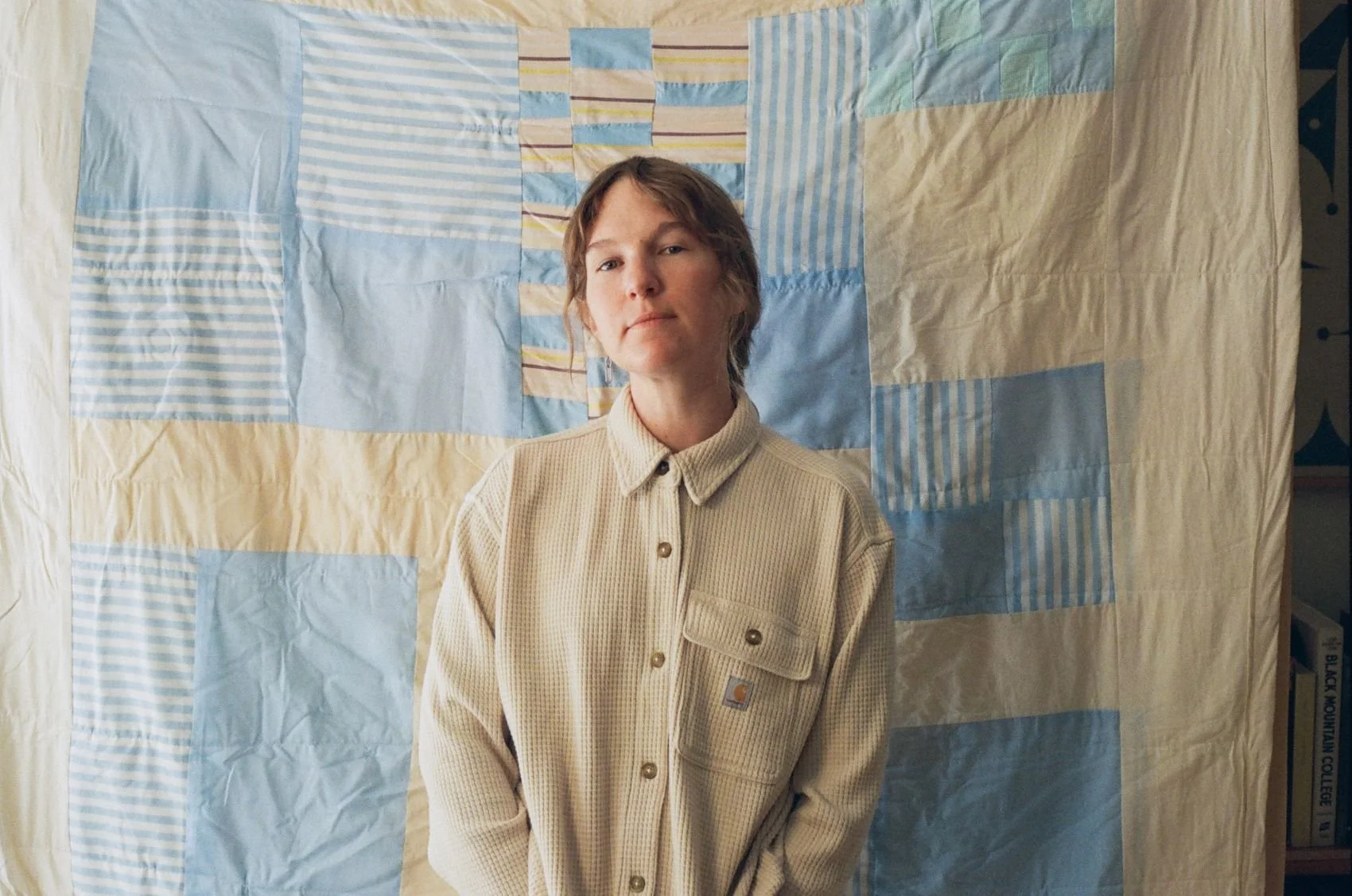 A person with short brown hair wearing a beige shirt stands in front of a patchwork quilt with various blue, yellow, and beige fabrics.