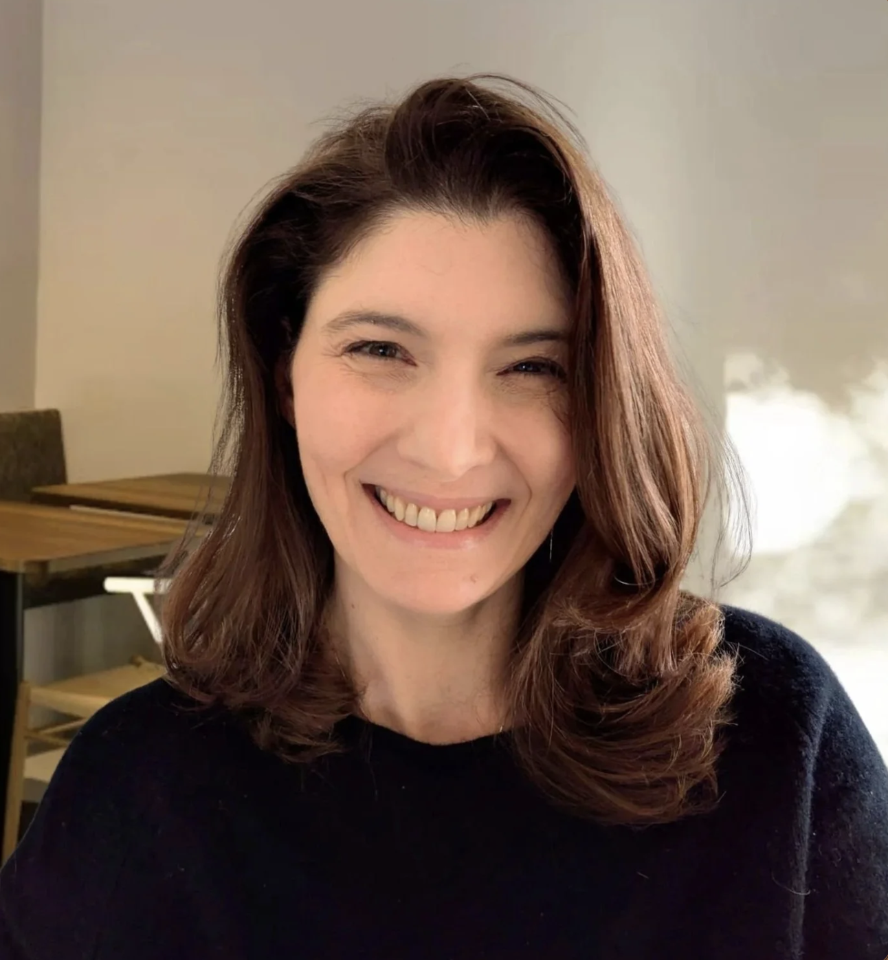 A woman with shoulder-length wavy brown hair smiling and showing her teeth, wearing a black top, with a wooden table and chairs in the background.