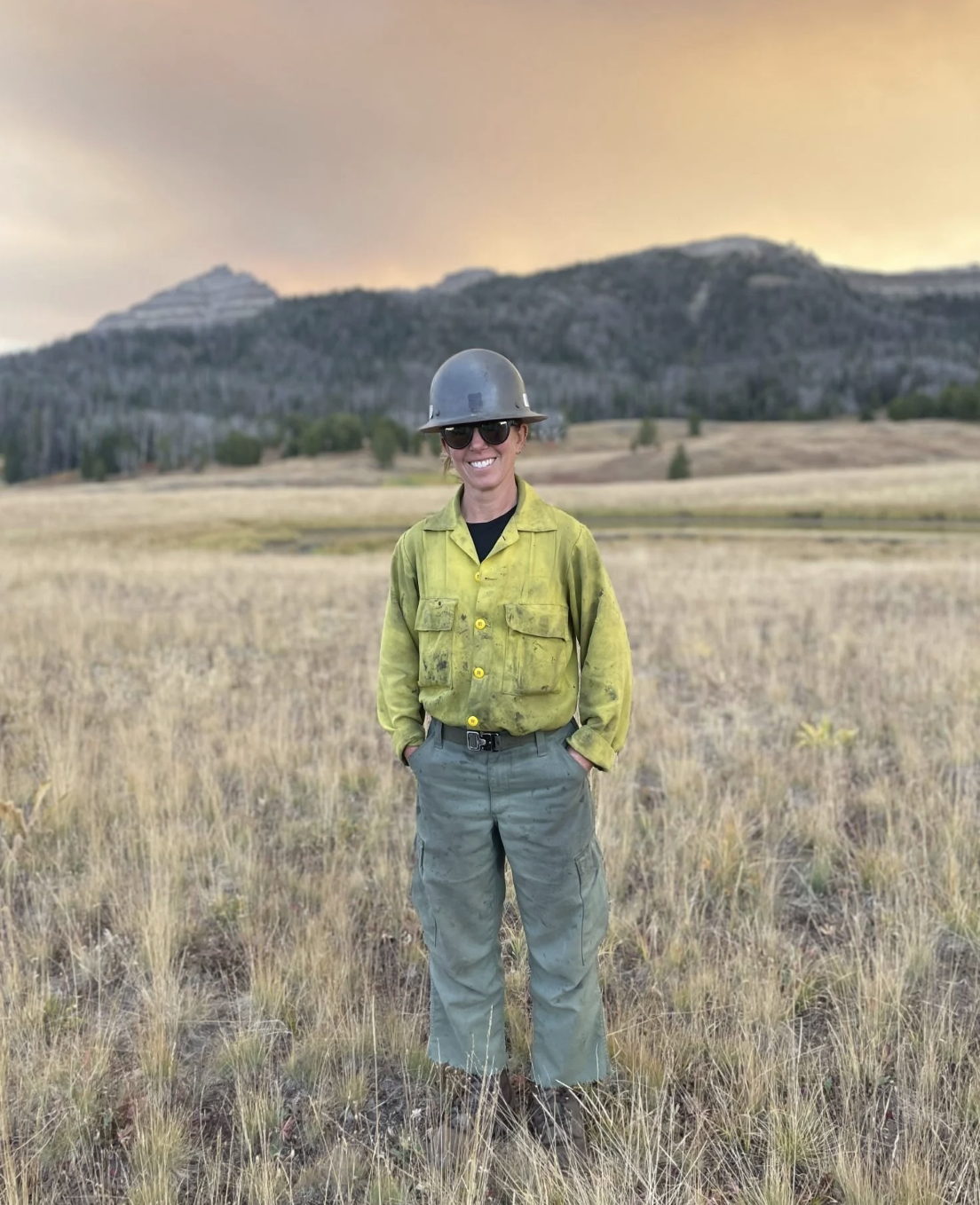 A woman wearing a green work shirt, gray pants, a gray safety helmet, and sunglasses standing in a field with dry grass and hills in the background at sunset.