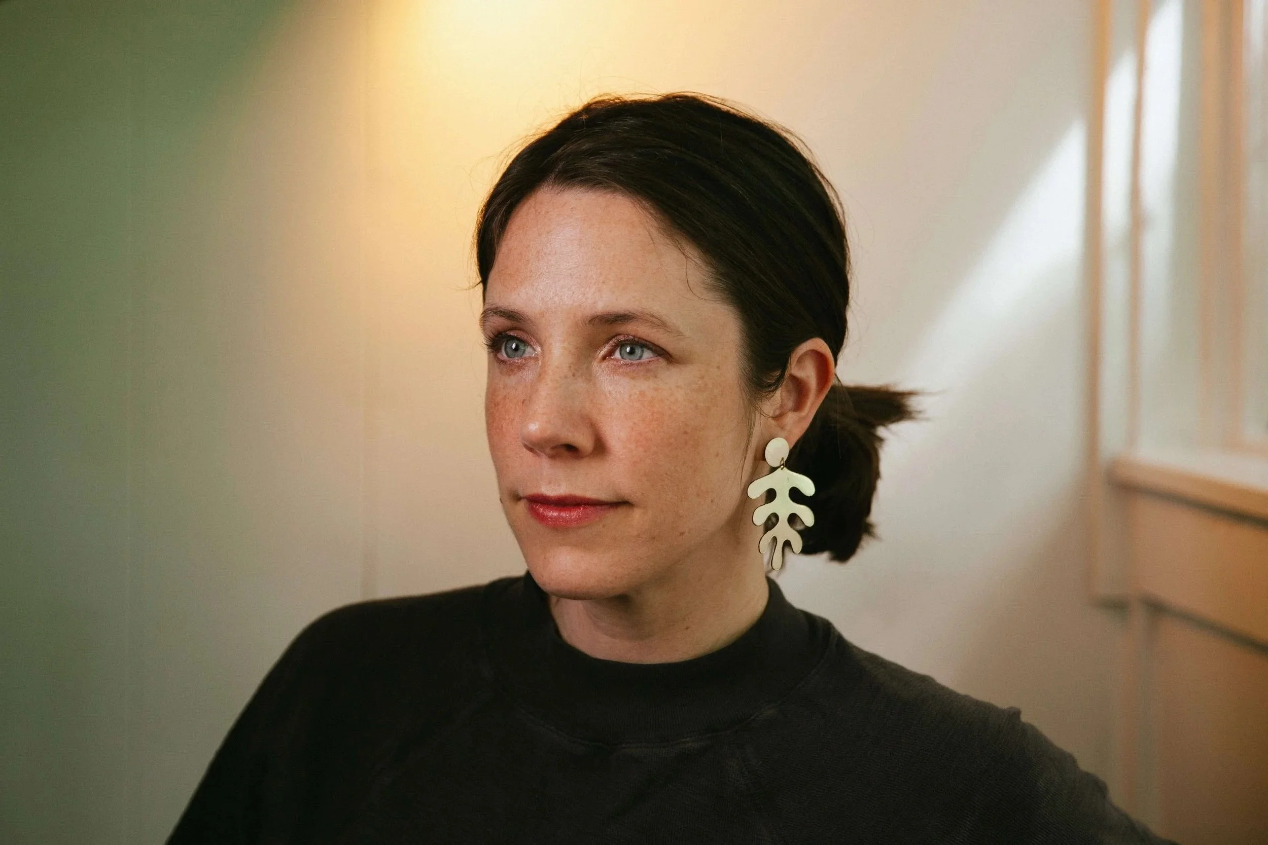Close-up of a woman with short dark hair, blue eyes, wearing a black top and statement earrings with leaf-shaped design, standing in front of a softly lit background.