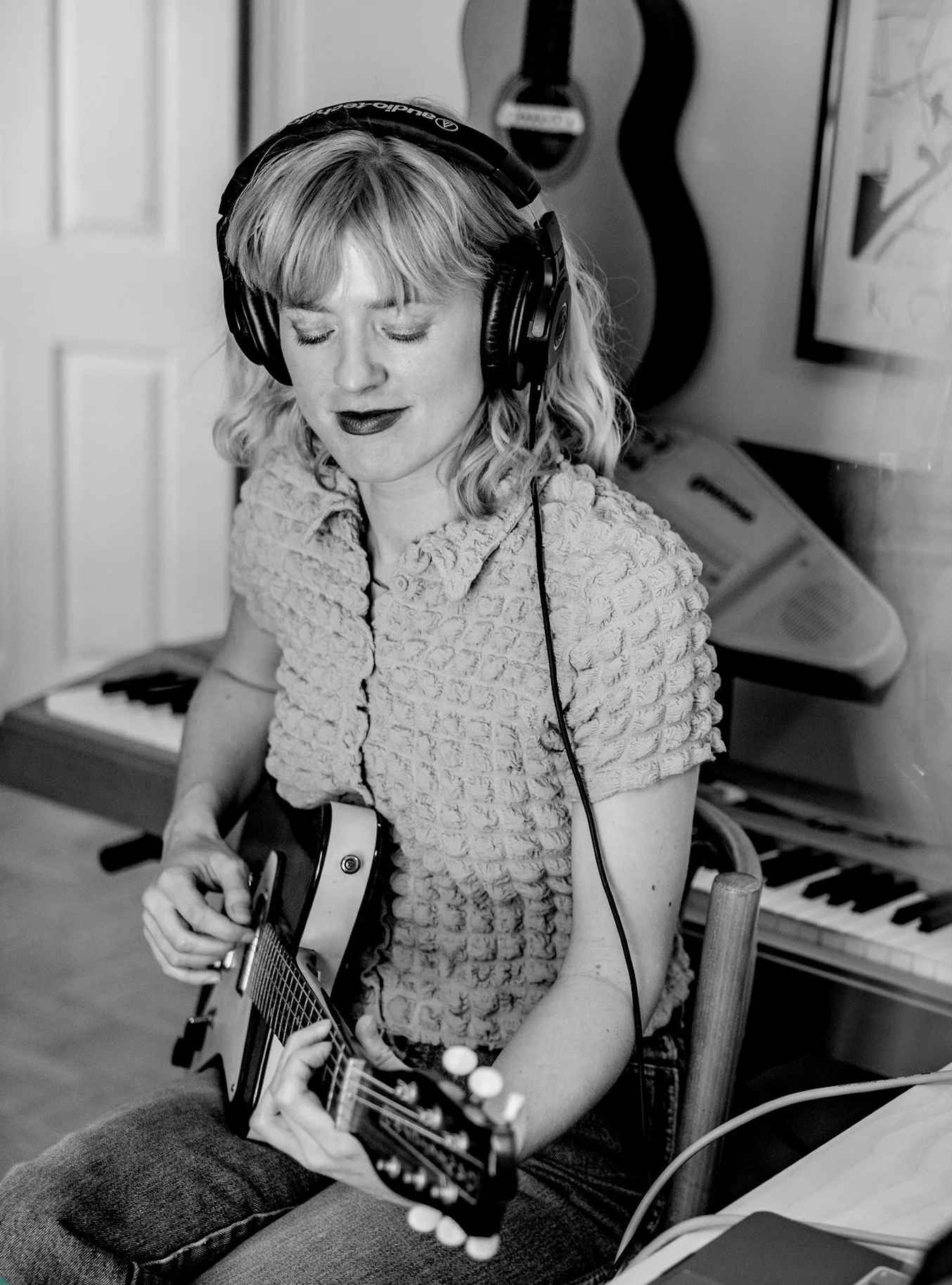 A woman with blonde hair wearing headphones playing an acoustic guitar in a room with musical instruments, including a keyboard and a guitar hanging on the wall.