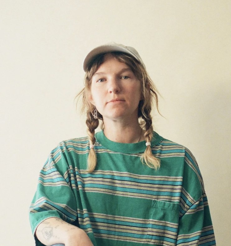 A person with braided hair, wearing a green striped t-shirt and a baseball cap, standing against a plain background.