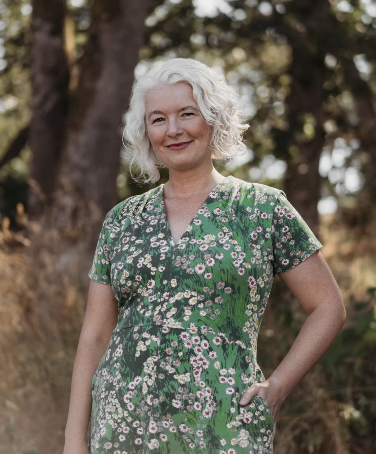 A woman with white curly hair wearing a green dress with white flowers, standing outdoors in front of trees, smiling gently.