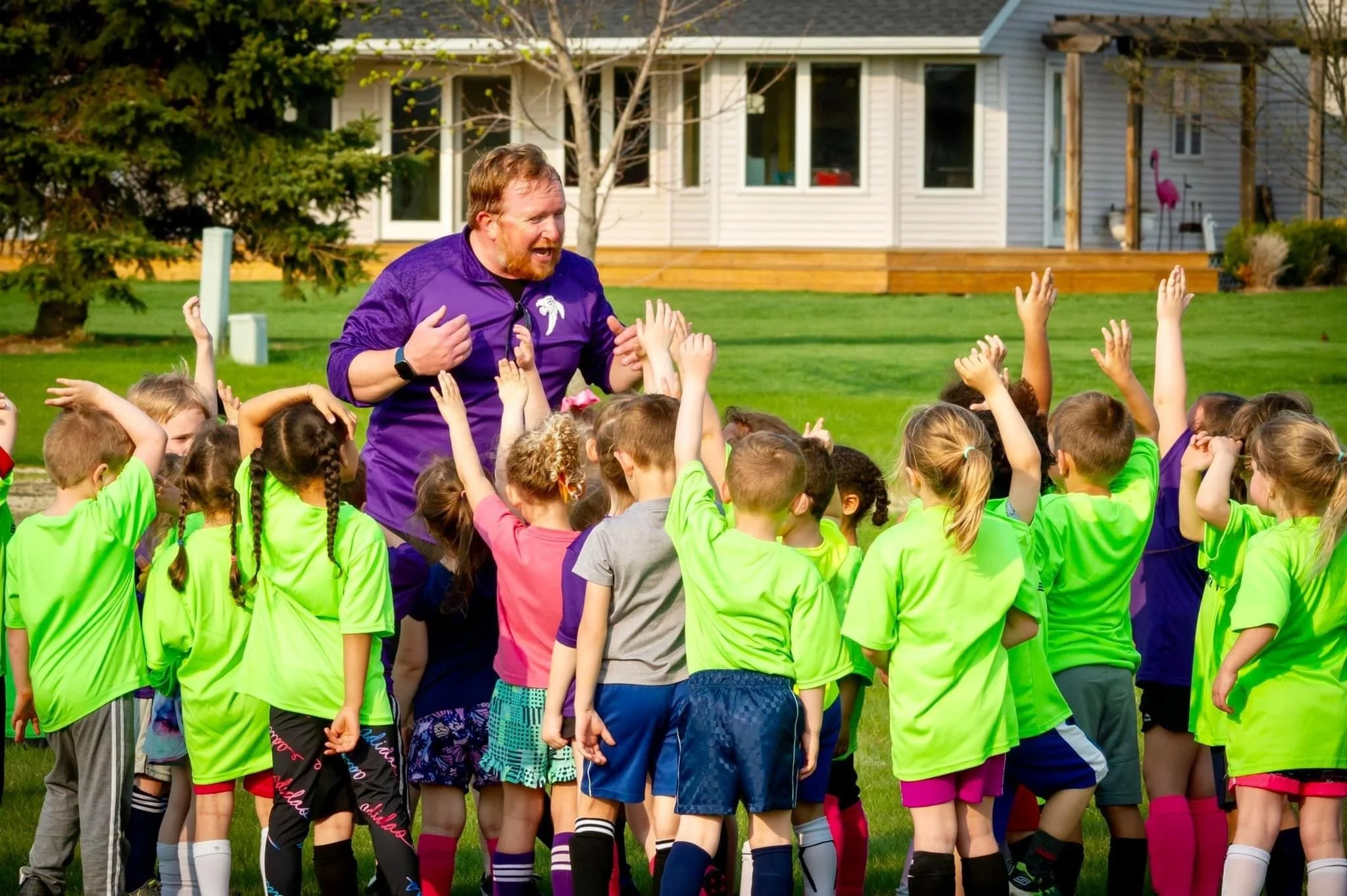 Christian giving instructions to a group of children at Sheboygan Falls FC on a grassy field with houses in the background.