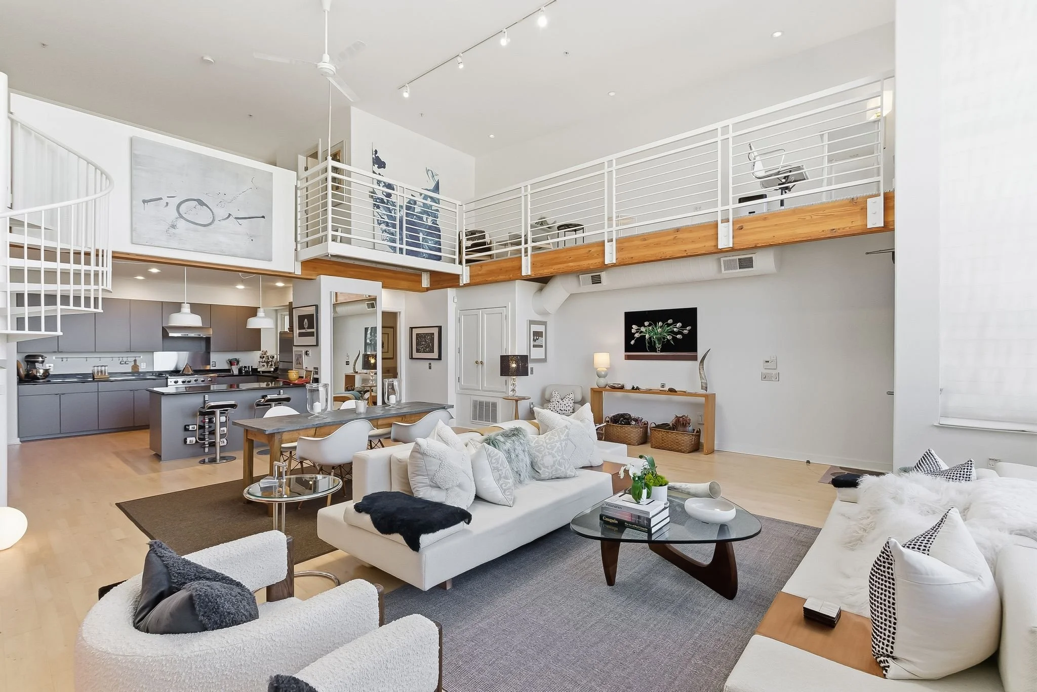 Spacious open-plan living room and kitchen area with a lofted workspace above, decorated in a modern minimalist style with white walls, light wood flooring, and contemporary furniture.