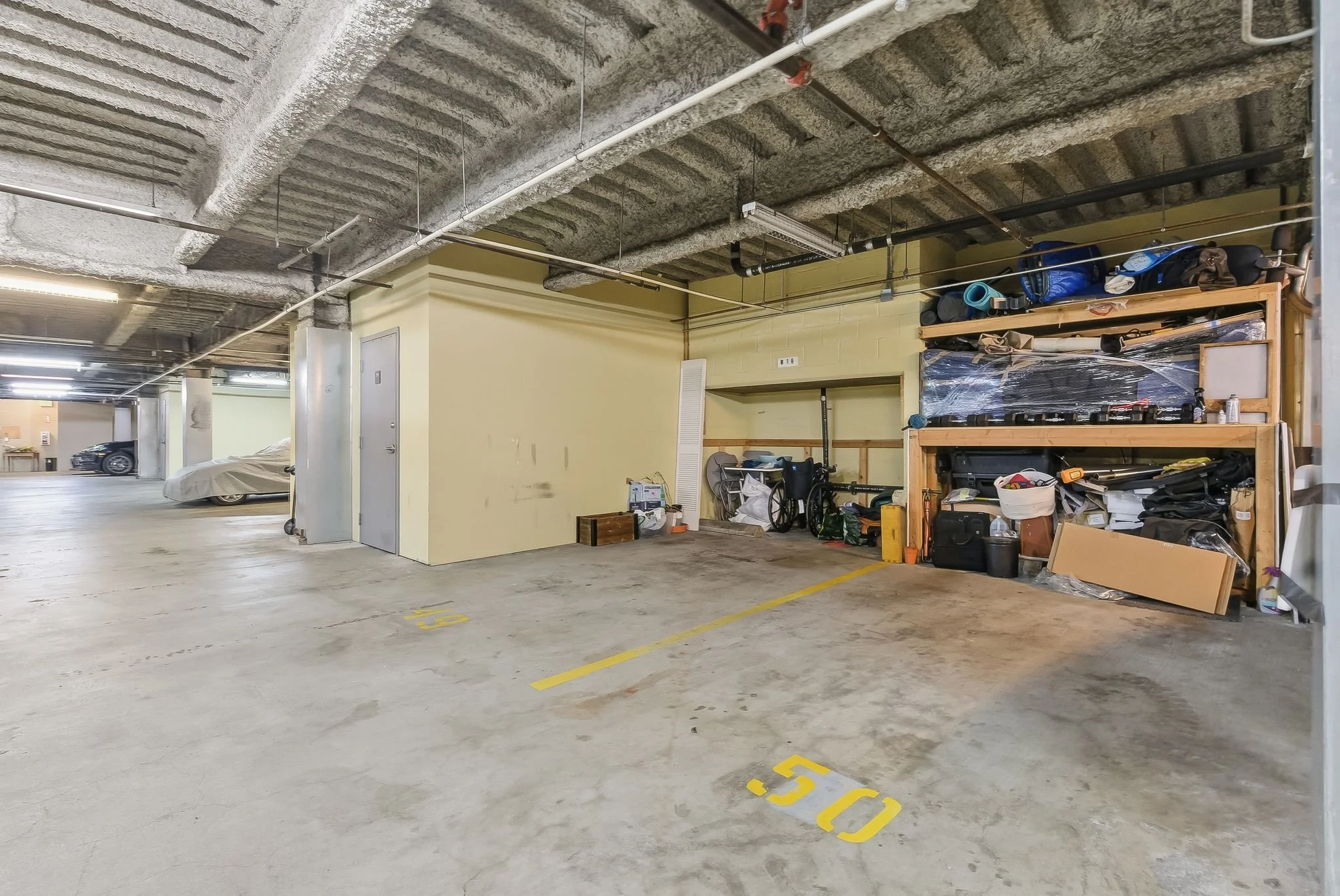 Underground parking garage with a lined parking space numbered 50, and a storage area with bins, bikes, and miscellaneous items on shelves.