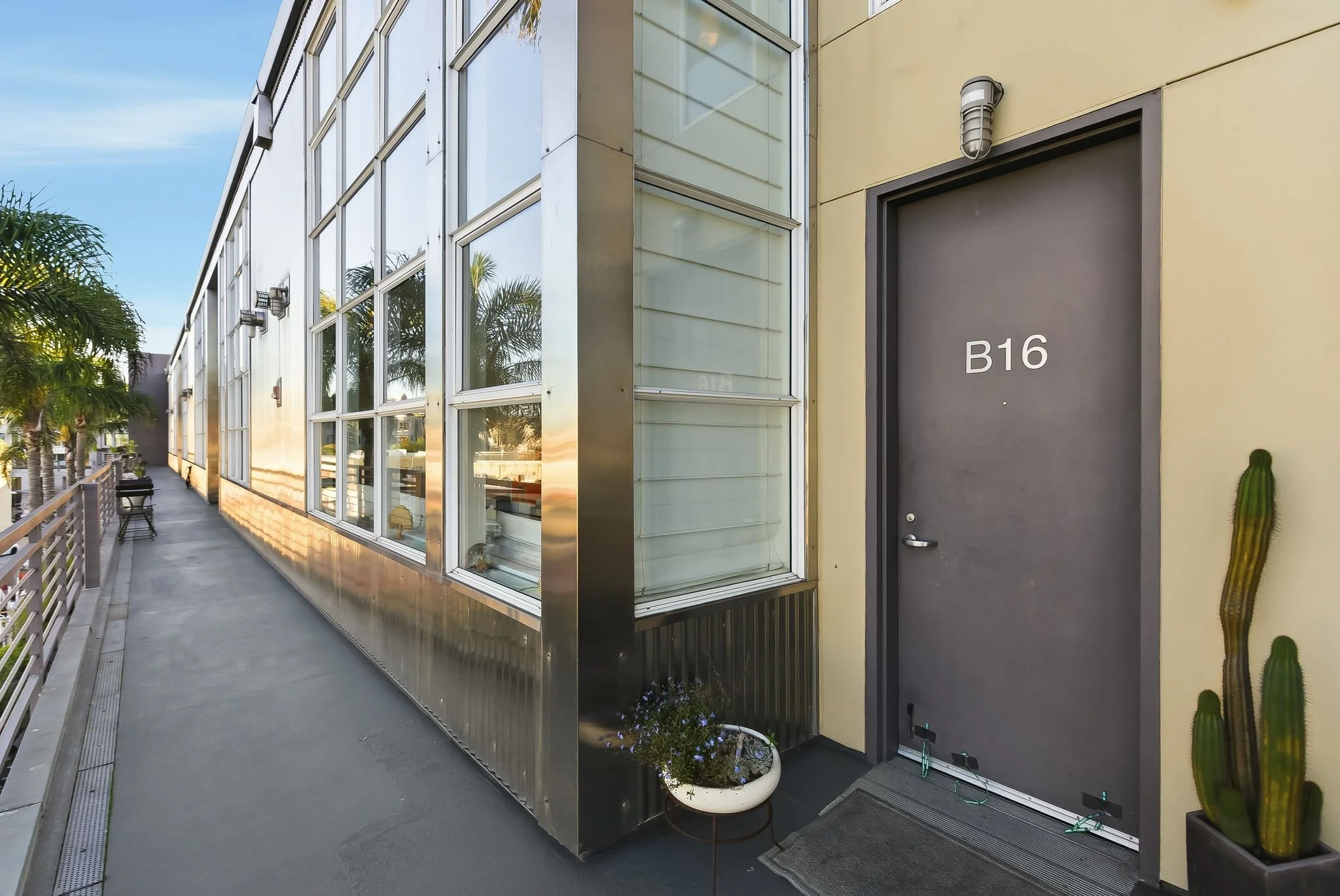 Exterior view of a modern building with large glass windows, a gray door labeled B16, a cactus plant, and a planter with flowers near the entrance, in a sunny outdoor setting.
