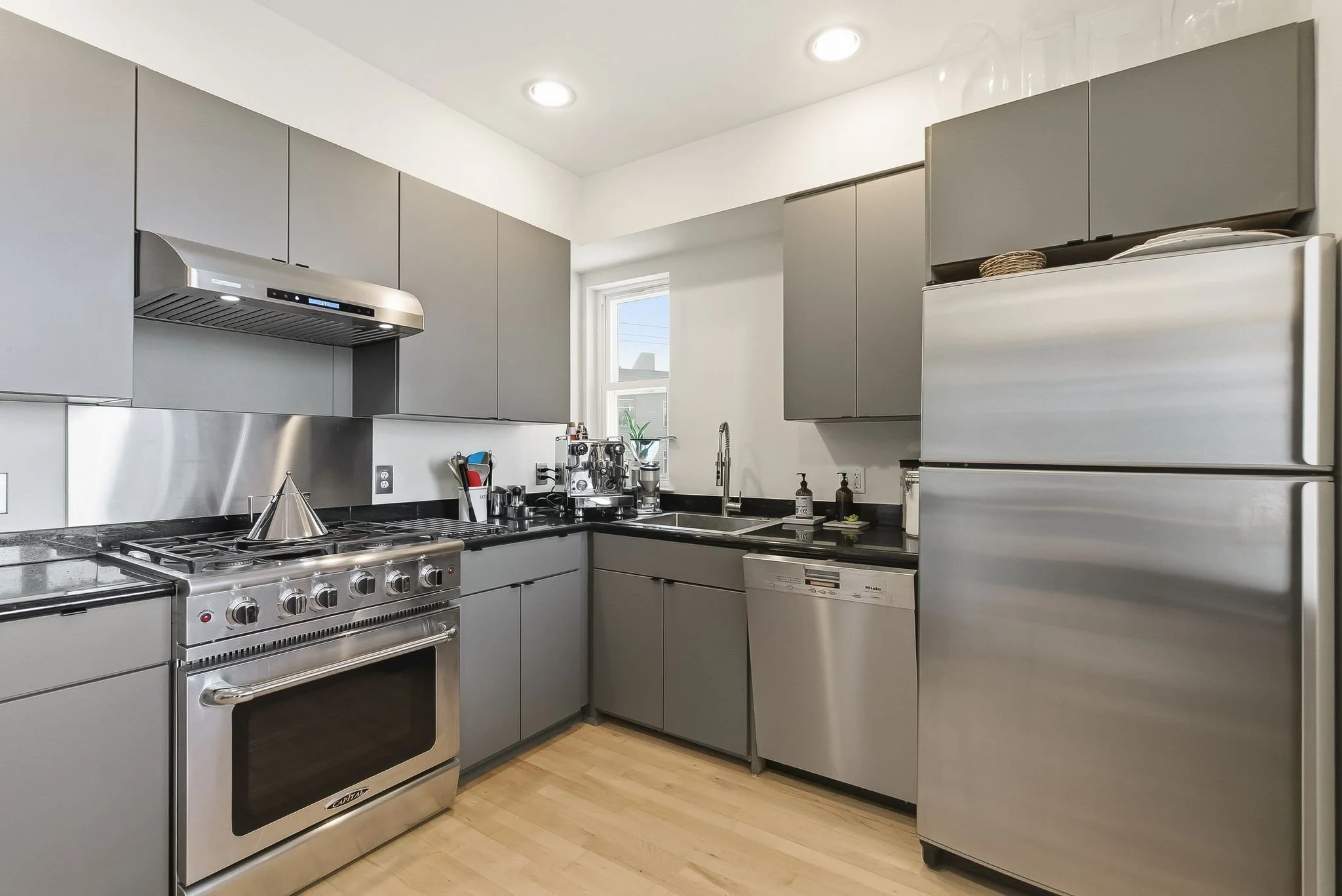 Modern kitchen with gray cabinets, stainless steel appliances including a refrigerator, oven, microwave, and dishwasher. Small window above the sink, black countertops, and hardwood flooring.