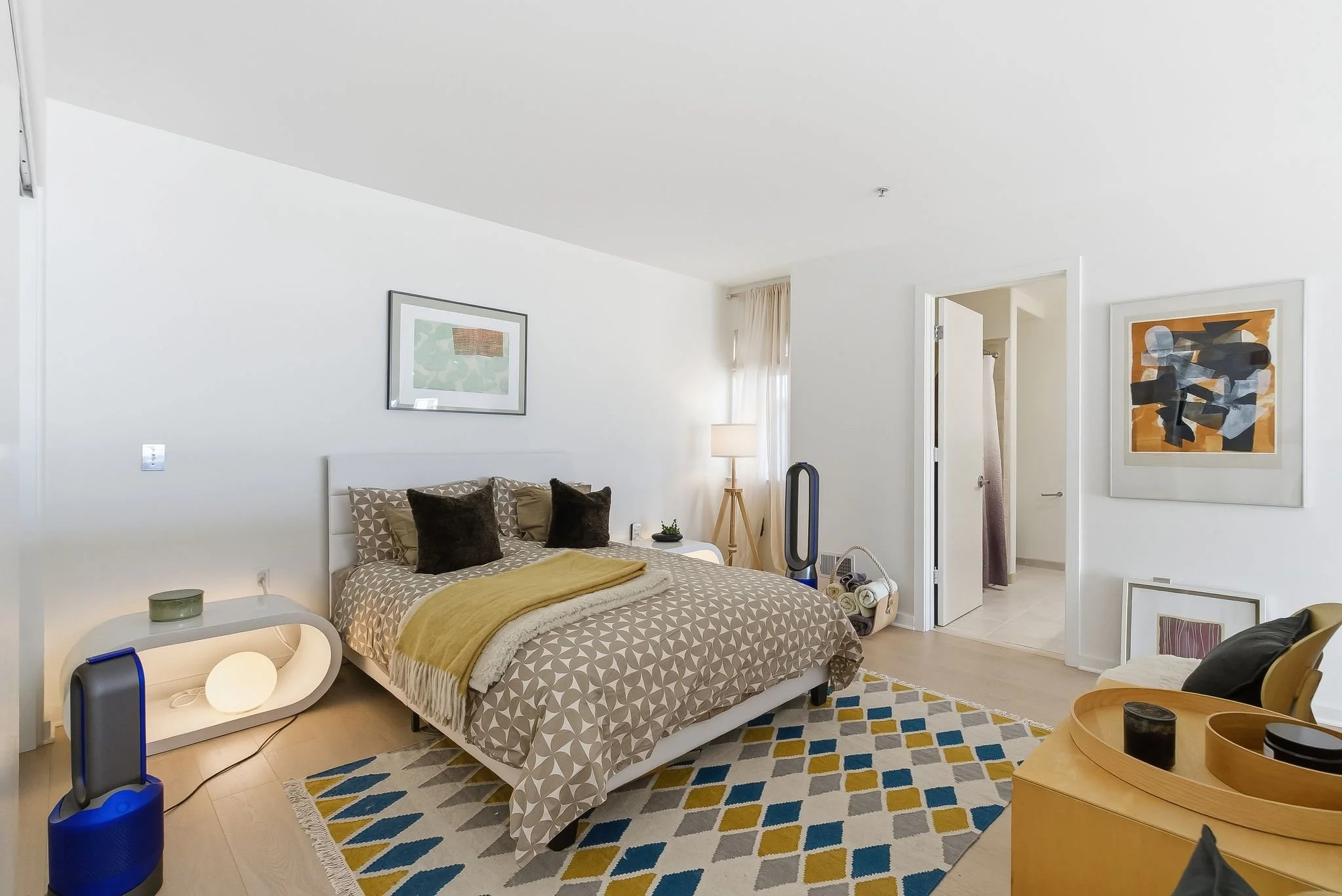 A cozy bedroom with a white bed, patterned bedding, and dark pillows. Side tables, artwork on the walls, a lamp, and a fan are visible. The room has light wooden flooring, a colorful geometric rug, and an open door leading to a bathroom.