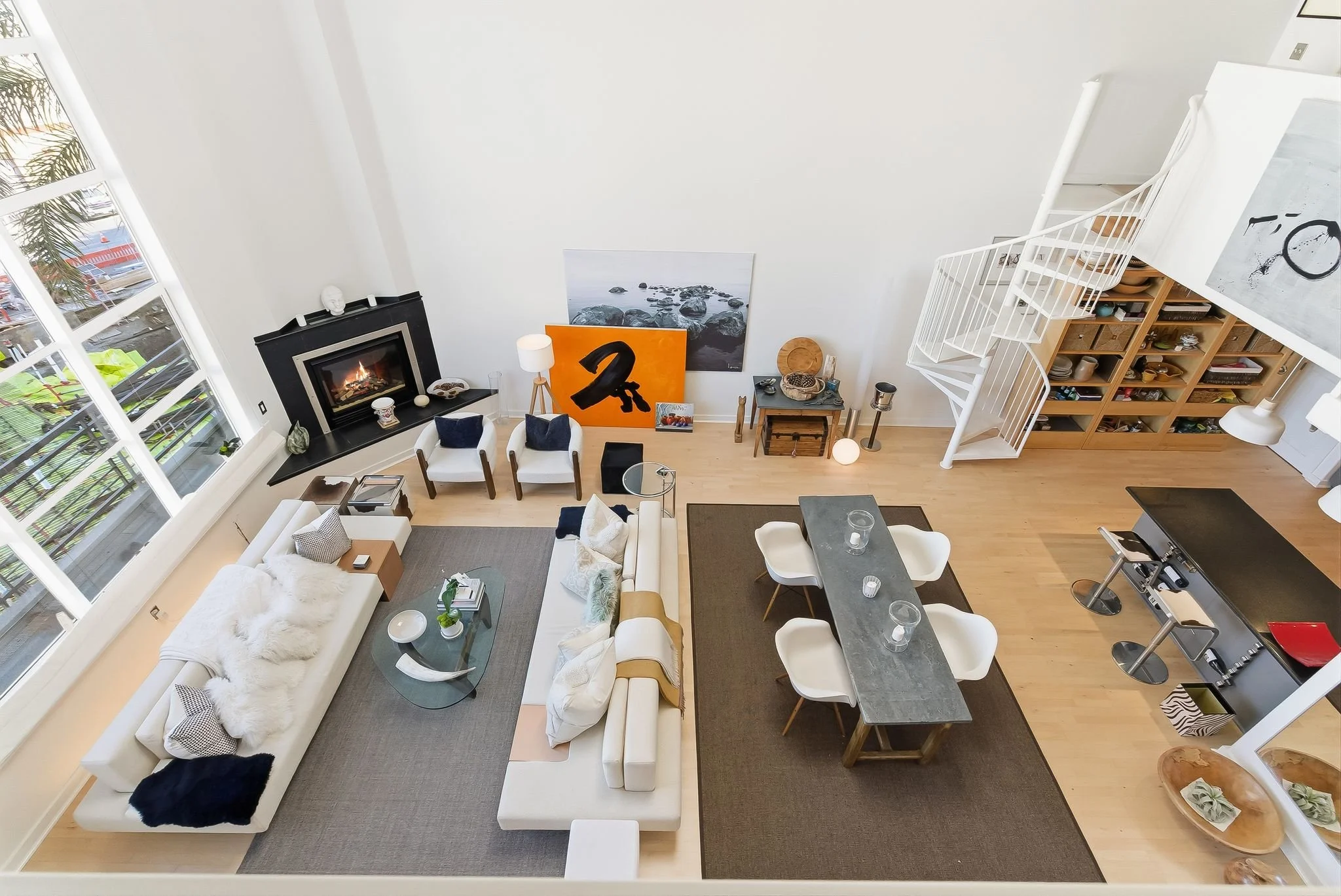 A spacious, modern living room with a white sofa, two white armchairs, a gray dining table with six white chairs, a fireplace, artwork leaning against the wall, and a wooden staircase leading to an upper level. Large windows on the left allow natural light to fill the room.
