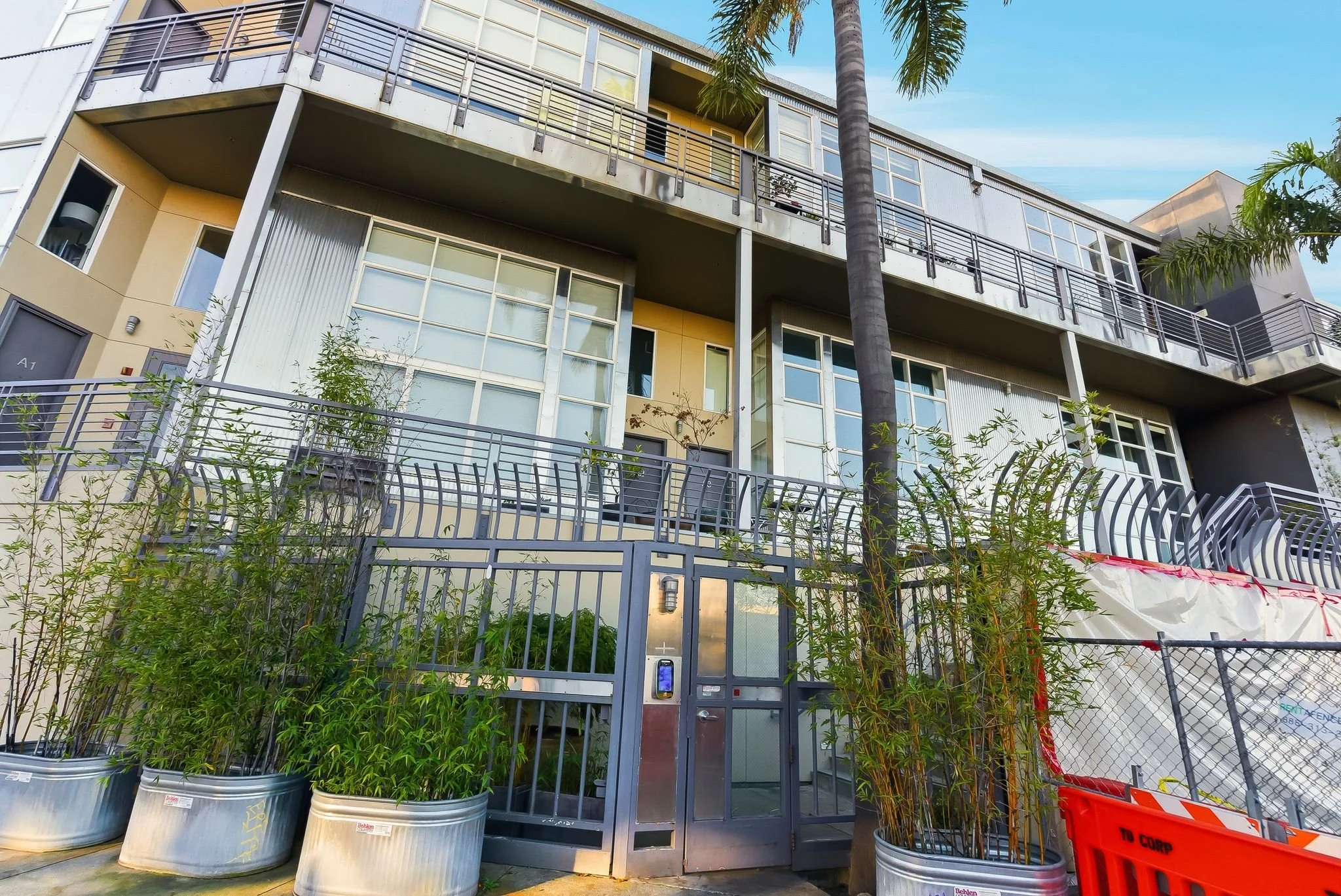 A modern multi-story residential building with large glass windows, metal balcony railings, and a gated entrance. There are several potted plants and bamboo along the fence, with some palm trees nearby. The sky is clear with some clouds.