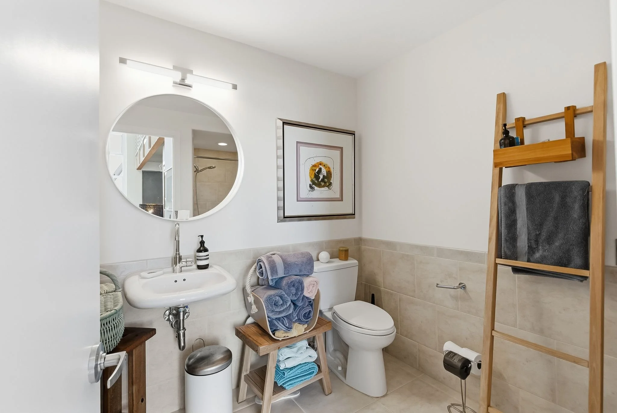 A clean bathroom with a round mirror above a small white sink, a framed abstract artwork on the wall, a wooden ladder-style towel rack holding a gray towel, and a toilet with a small stool filled with rolled towels on top. There are decorative items 