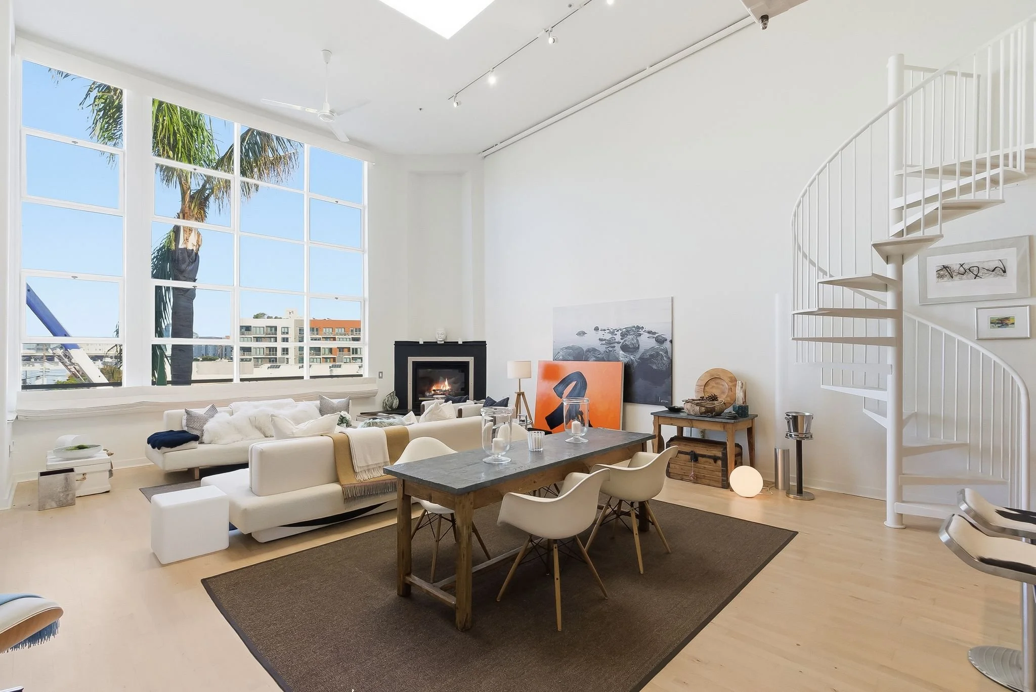 Bright modern living room with large windows, white walls, a fireplace, a large white sectional sofa, a wooden dining table with four white chairs, artwork leaning against the wall, and a spiral staircase in the corner.