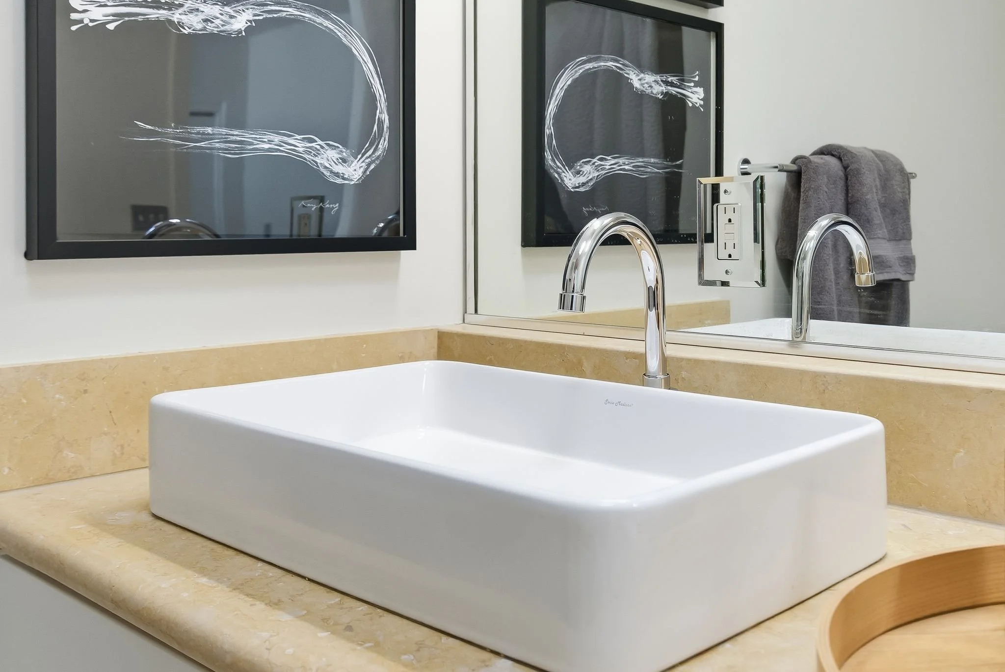 Bathroom sink with a modern rectangular white vessel basin, chrome faucet, beige countertop, large mirror, artwork with abstract white lines on black background, electrical outlet with cover, gray towel hanging on a towel rack.
