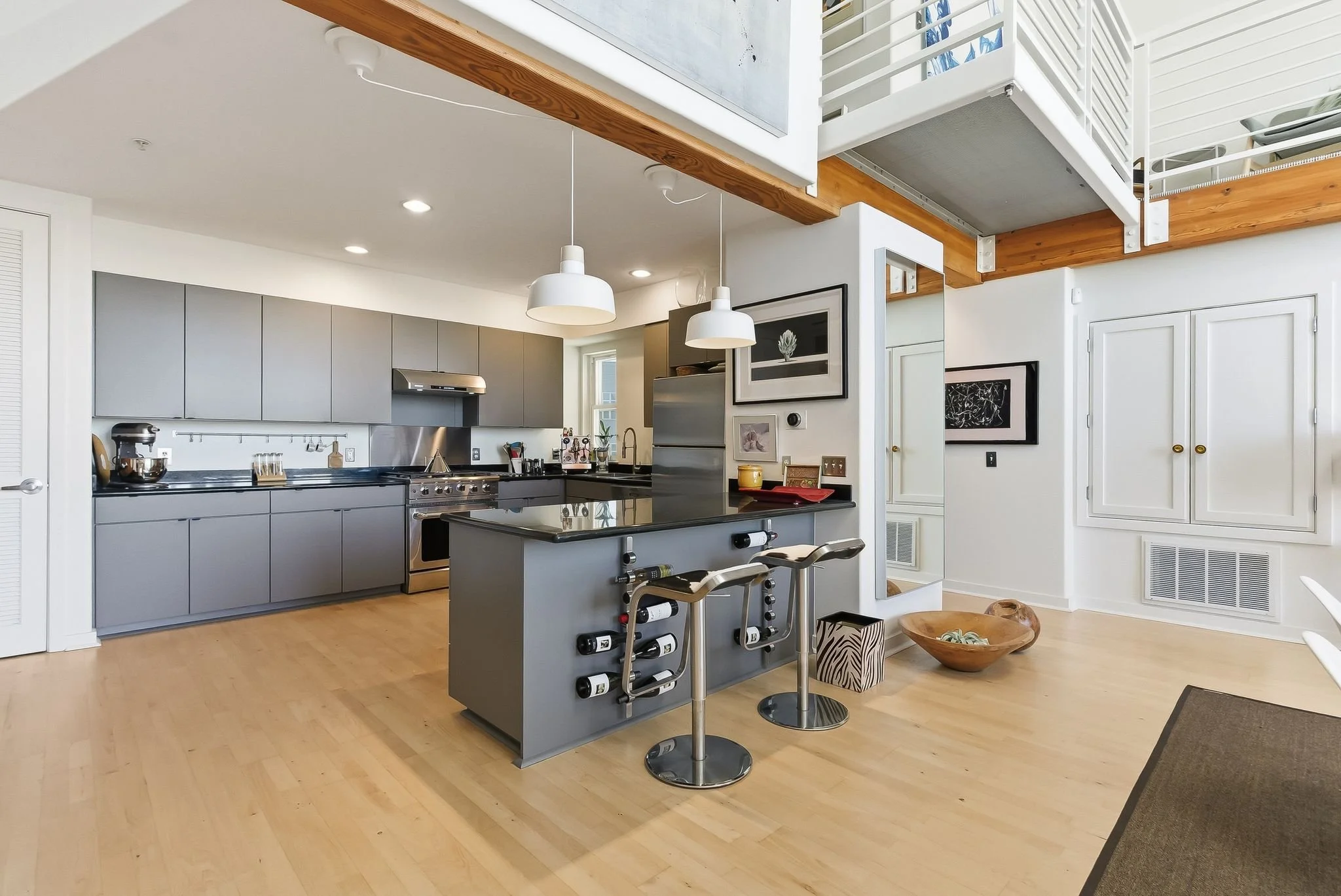 Modern open-concept kitchen with gray cabinets, stainless steel appliances, light wood flooring, and a gray island with wine bottle holders and two bar stools.