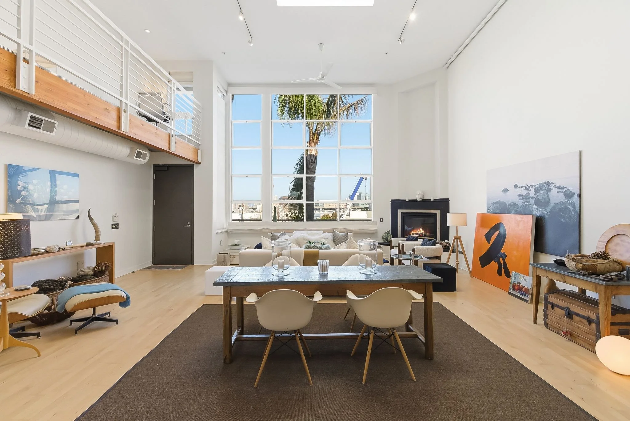 Modern living room with large window, white walls, wooden flooring, and contemporary furniture, including a dining table with chairs, a sofa, armchairs, a fireplace, and art decorations.