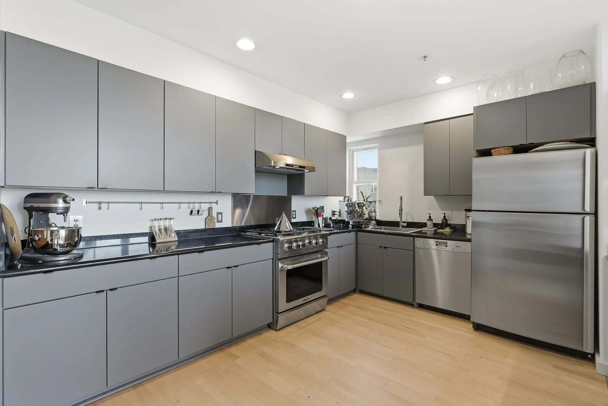 Modern kitchen with gray cabinets, black countertops, stainless steel appliances including a refrigerator, oven, and a dishwasher, wooden flooring, and a window letting in natural light.
