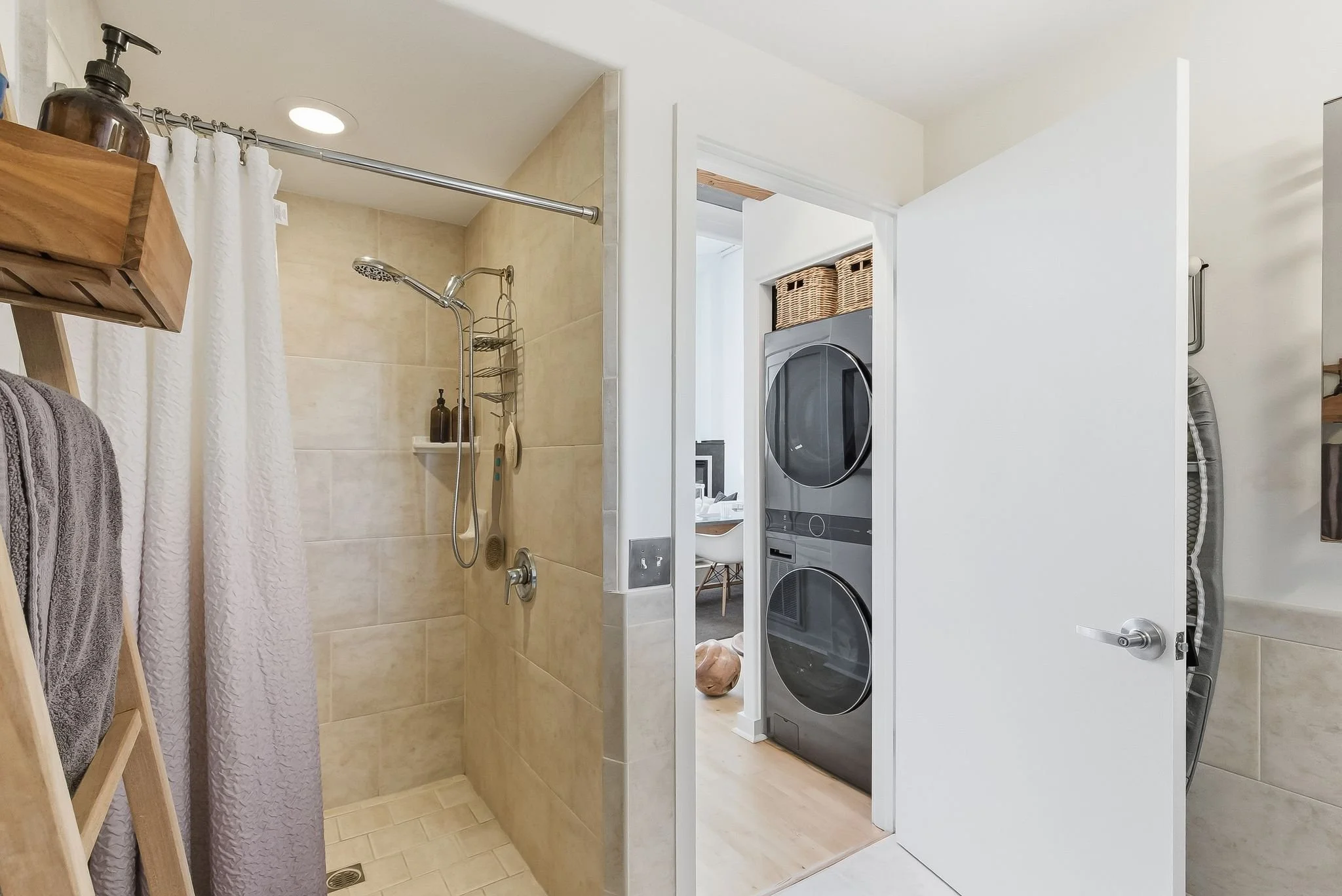Bathroom with shower area, beige tiles, shower shelves, and laundry area with stacked washer and dryer behind a white door.