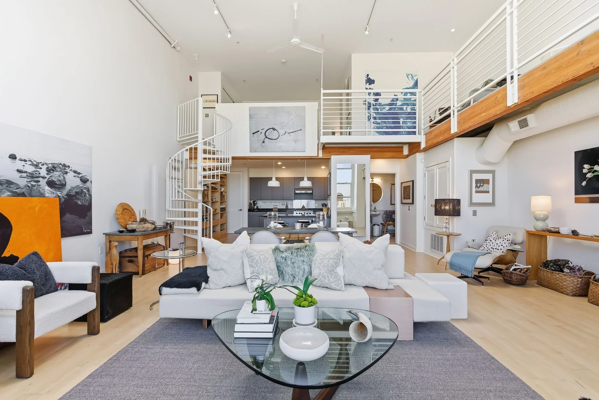 Bright living room with white sofa, glass coffee table, and decorative pillows, overlooking a modern kitchen in an open-concept space with hardwood floors and a loft area.