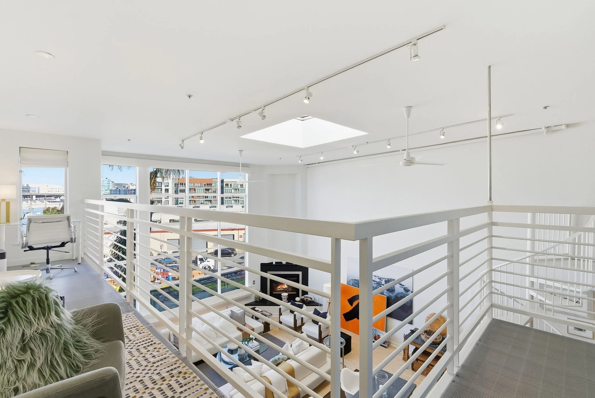 A modern, open-plan living room in a high-rise apartment with white walls, large windows, and a view of city buildings. The space features a white railing, a ceiling fan, a flat-screen TV, a fireplace, and contemporary furniture.