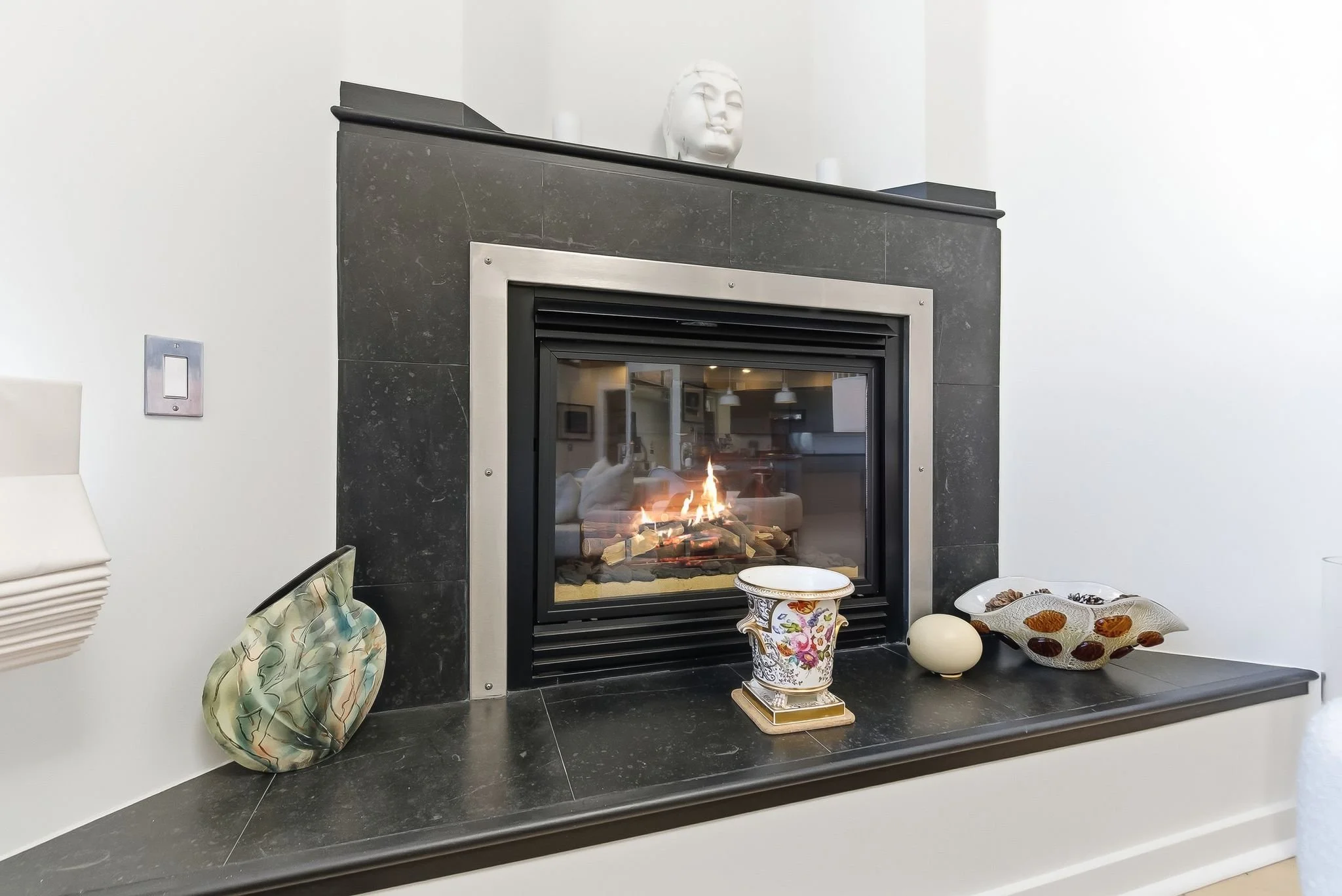 A modern gas fireplace surrounded by black stone with various decorative objects on the mantel and shelf, including ceramic vases and bowls, with a reflection of a living room in the glass.