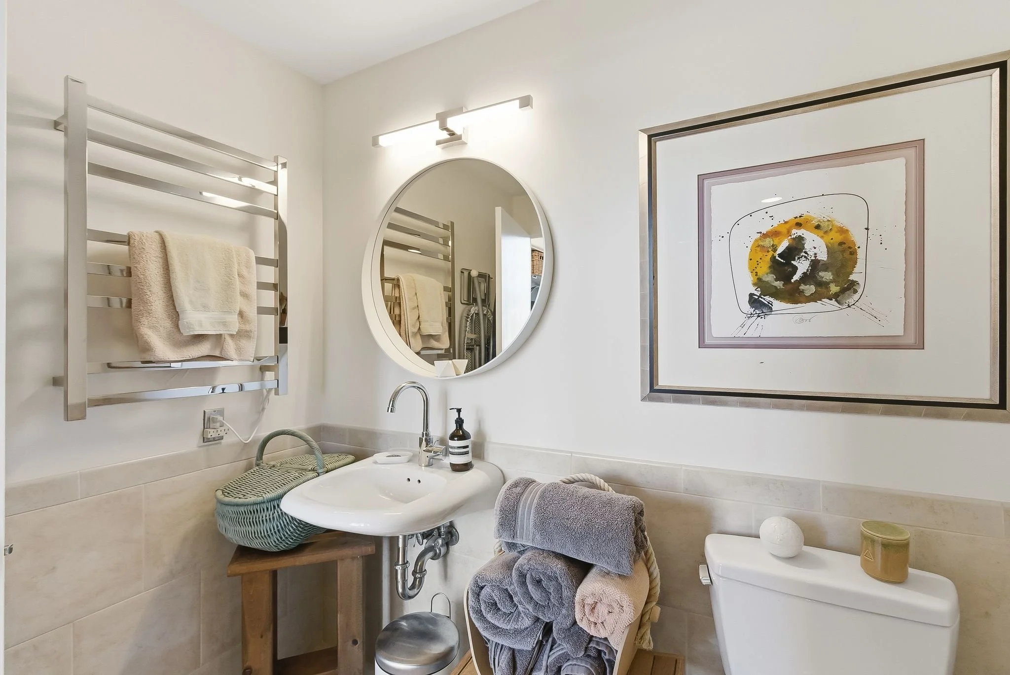 A bathroom with a white sink, mirror, heated towel rack, abstract artwork, and neatly folded towels.