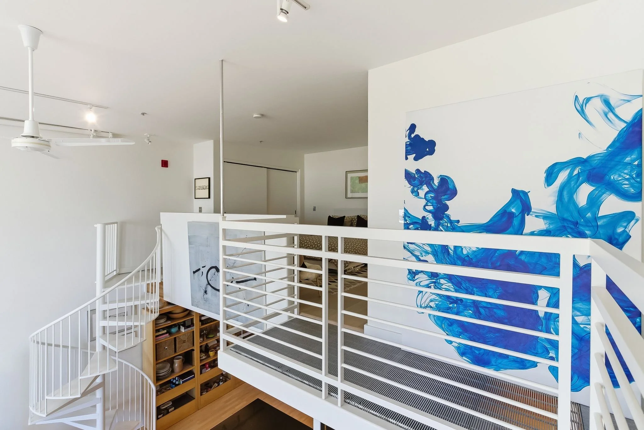 Modern interior of a two-story home with a white staircase, a large blue abstract ink splash mural on the wall, and a partial view of a living room with beige chairs and framed art.