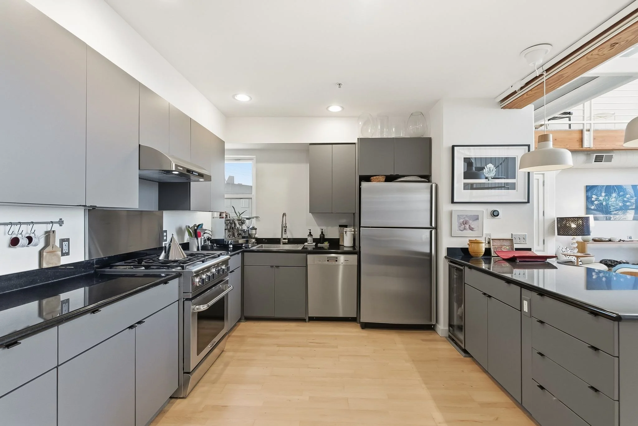 Modern kitchen with gray cabinets, stainless steel appliances, black countertops, light wood flooring, and artwork on the walls.