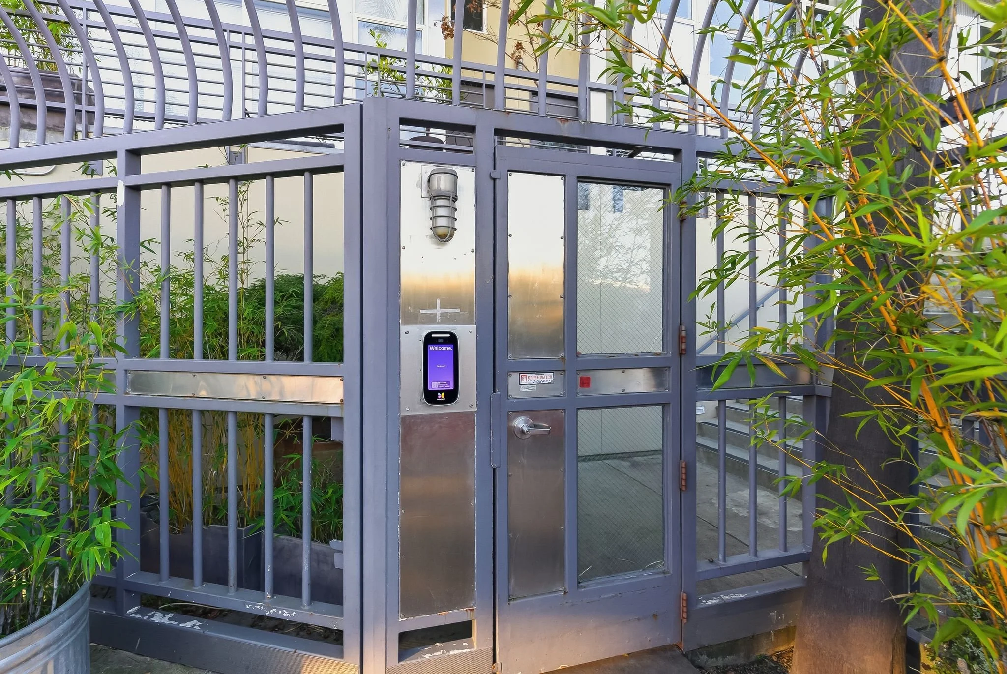 A gated outdoor enclosure with a metal structure, containing plants inside. There is a glass door with a metal frame and a digital keypad or access panel on the side, along with a light fixture on top.