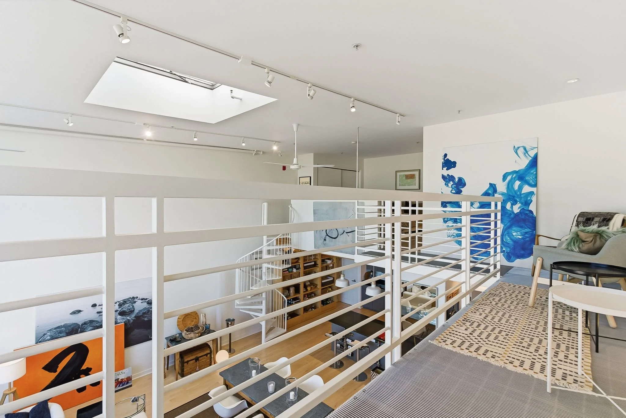 Interior view of a modern, minimalistic apartment with white walls, a skylight, and artwork. A balcony overlooks a living space below with a black table, chairs, and shelves.
