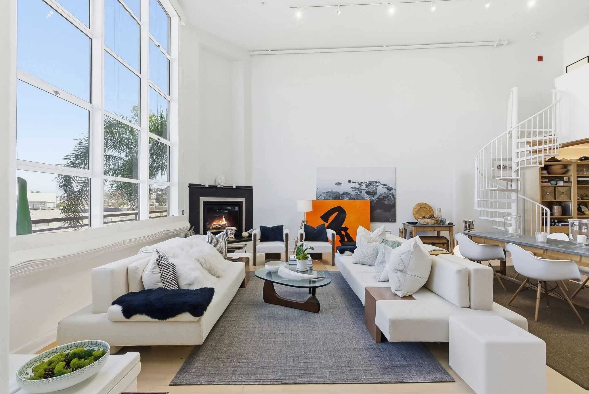 Modern living room with high ceiling and large windows, white sofas with pillows, a coffee table with decor, and artworks on the wall, with a staircase on the right.