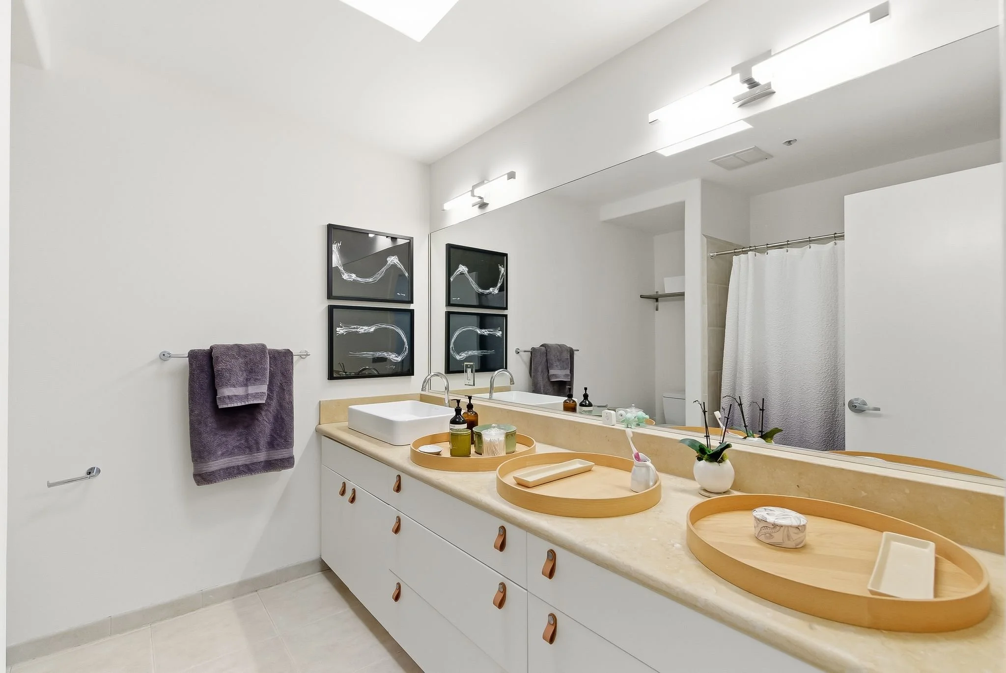 Modern bathroom with white walls, large mirror, beige countertop, white sink, black framed artwork, gray towels, wooden trays with toiletries, small plant, toothbrush holder, shower with white curtain.