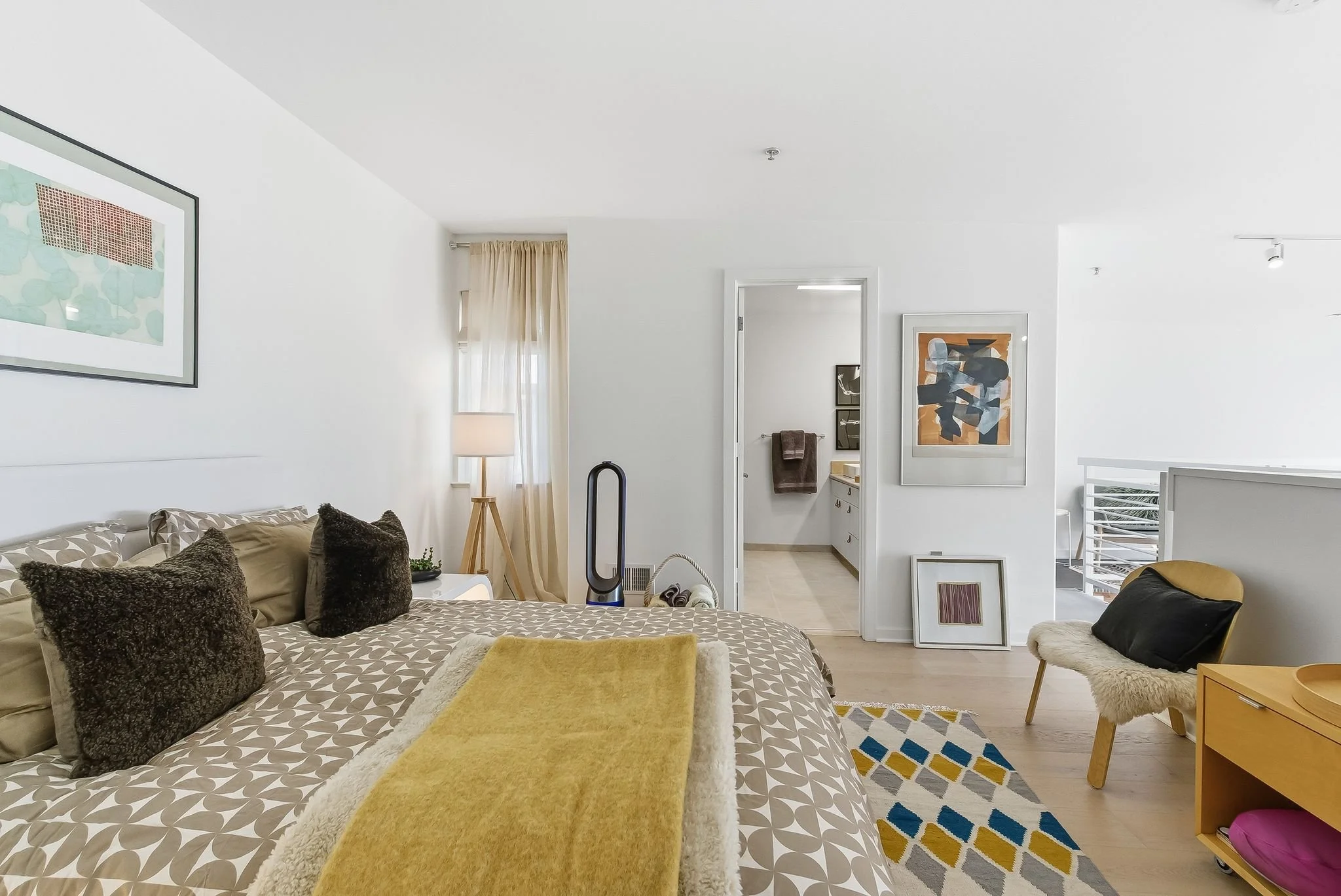 A modern bedroom with a bed covered in a patterned duvet and pillows, a yellow throw blanket, a bedside lamp, artwork on the walls, a beige chair with a black pillow, a wood side table, a colorful rug, and a Dyson bladeless fan, with an open door lea