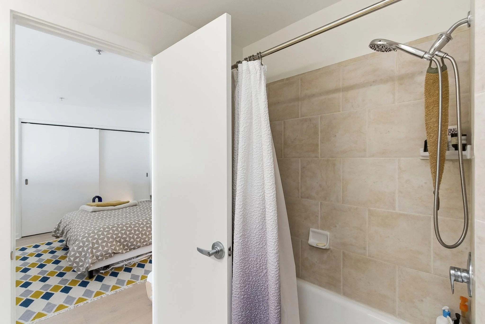 Bathroom with shower and bathtub, partially open door leading to bedroom with bed, patterned bedding, and a colorful geometric rug.