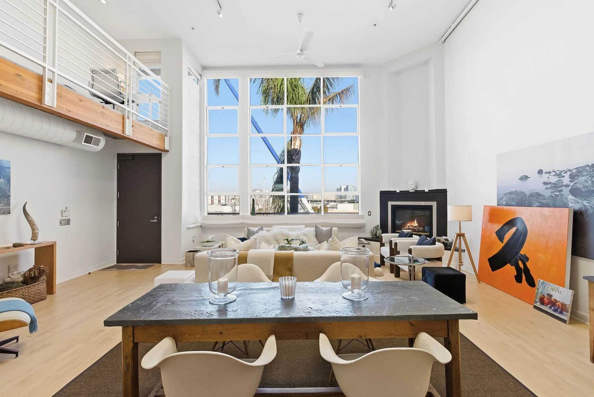 Bright, modern living room with large windows showing a palm tree outside. Features include a dining table with chairs, a fireplace, artwork, and various seating options.