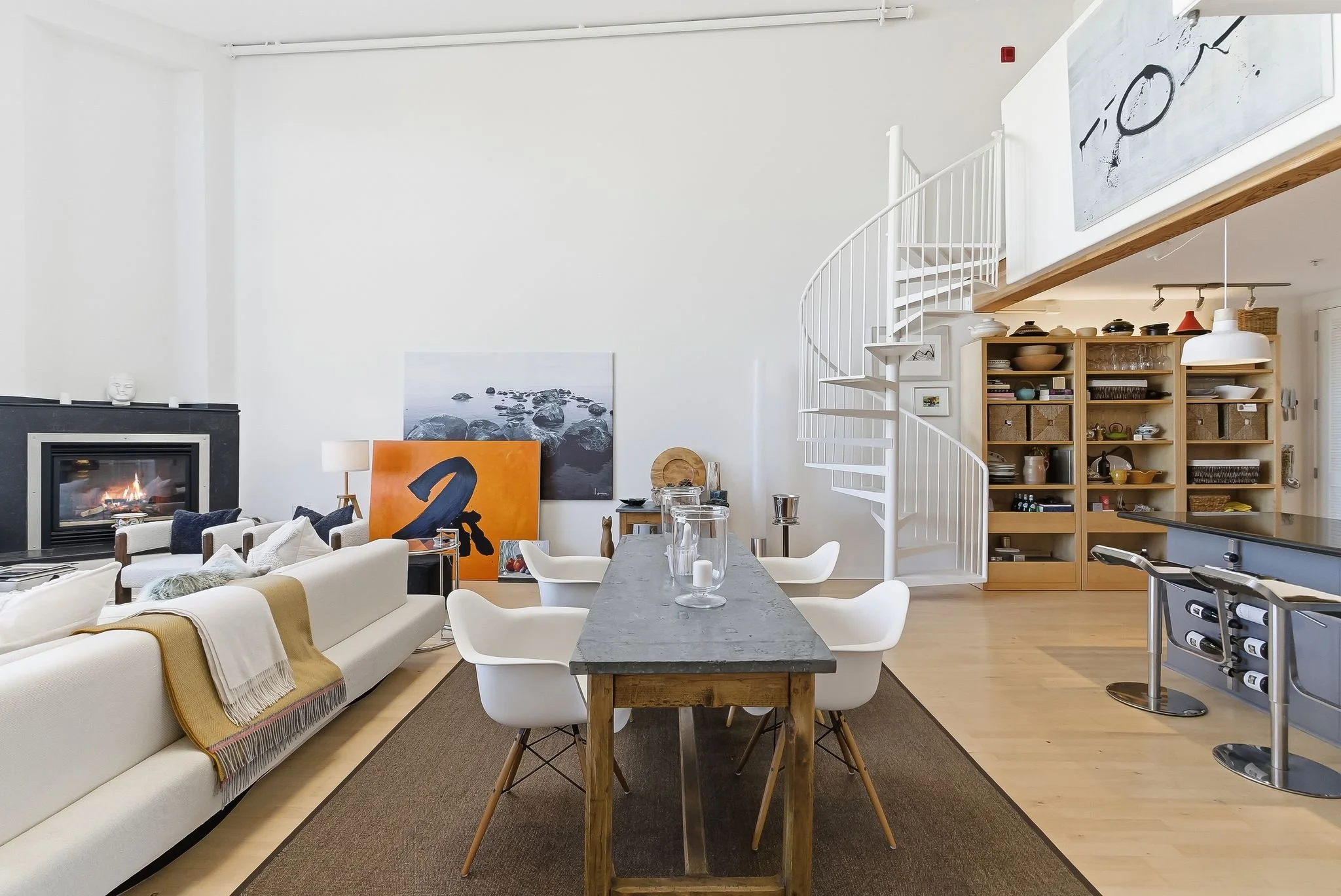Modern open-concept living and dining area with a fireplace, spiral staircase, and a wooden shelving unit filled with dishes and decor.
