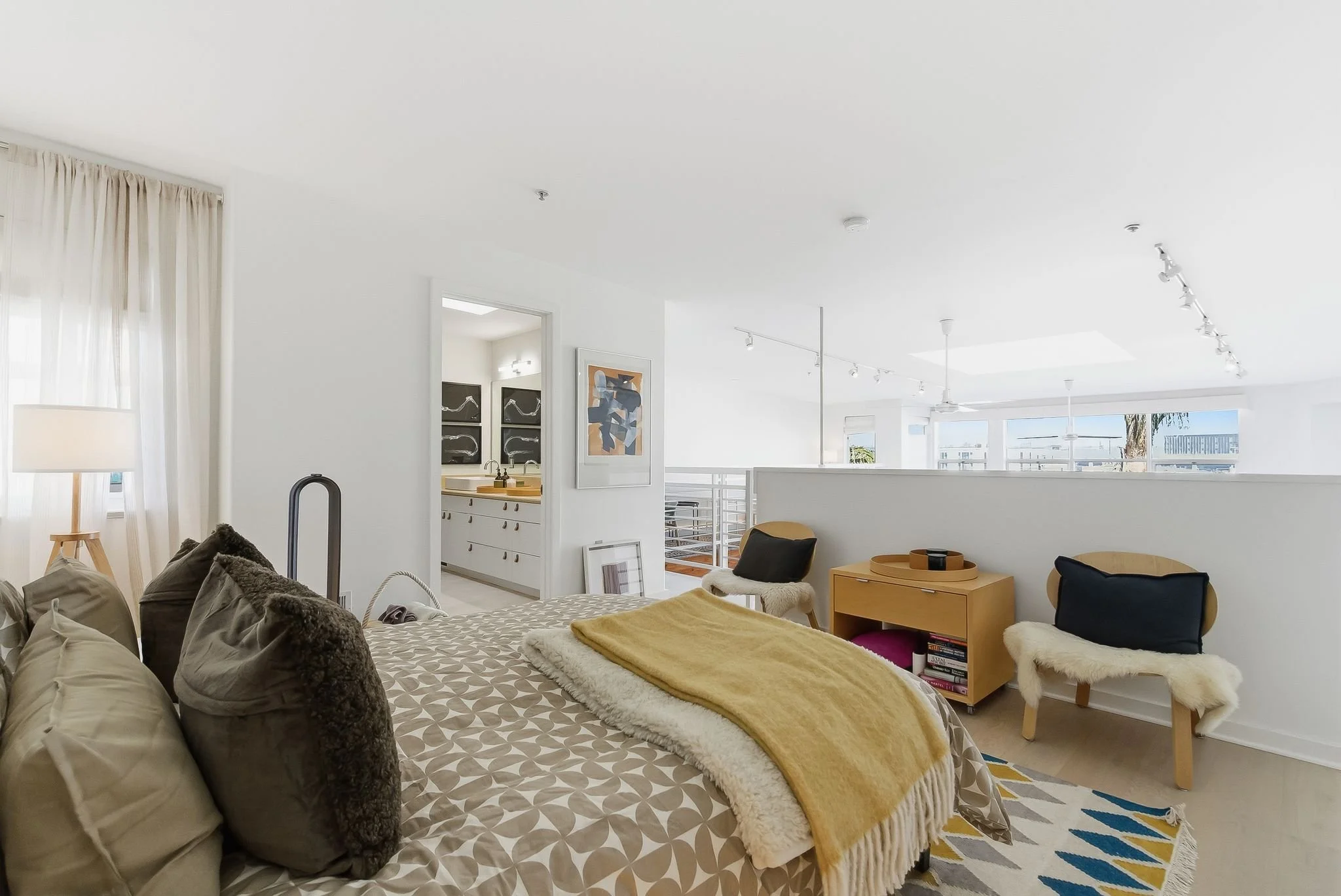 A bright, modern bedroom with white walls and a large bed covered in beige and brown pillows, a patterned bedspread, and a yellow throw blanket. There are two chairs with black and fur cushions, a wooden side table with books, and a colorful rug on t
