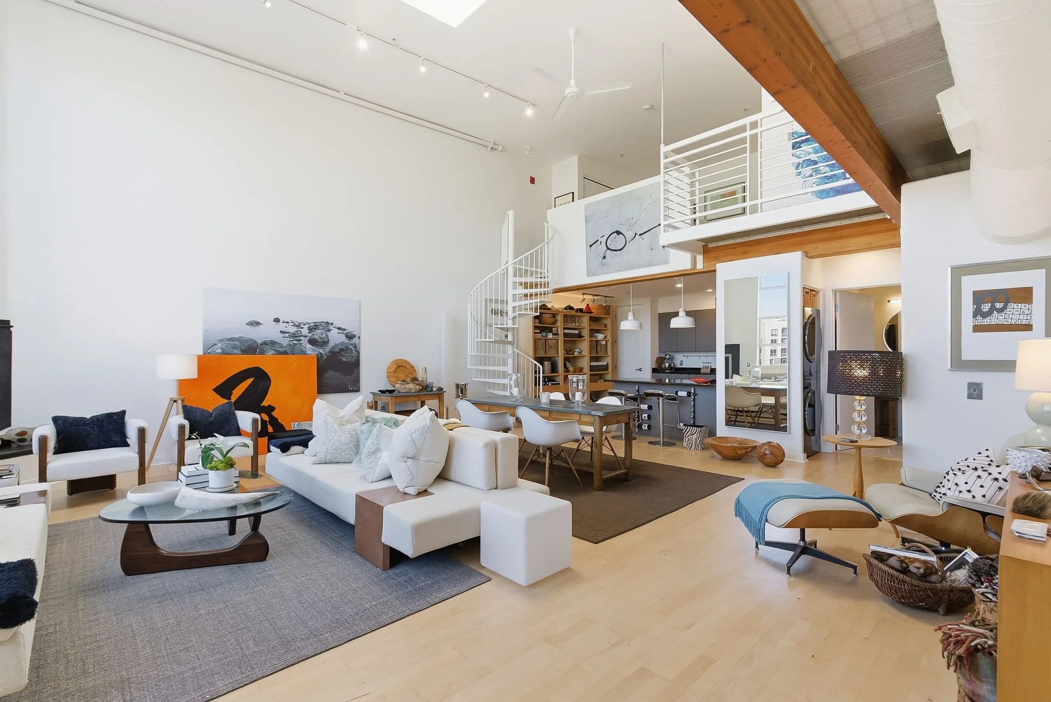 A spacious, modern living room with white walls, hardwood floors, and high ceilings. It has a white couch, a coffee table, and various chairs, with artwork on the walls. An open kitchen with barstools is visible in the background, and a spiral stairc