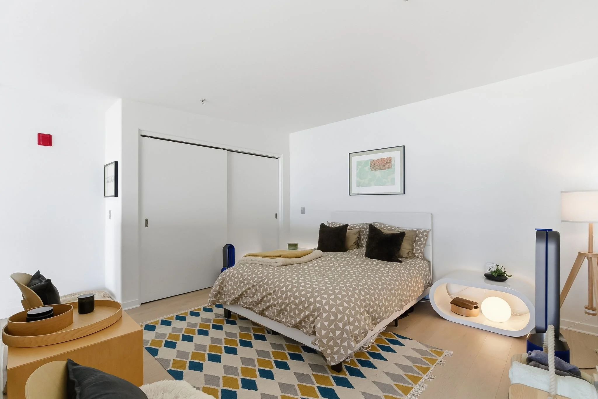 Modern bedroom with white walls, a bed with geometric-patterned bedding, and black and beige pillows. There is a patterned rug on the floor, a nightstand with a lamp, and artwork on the walls.