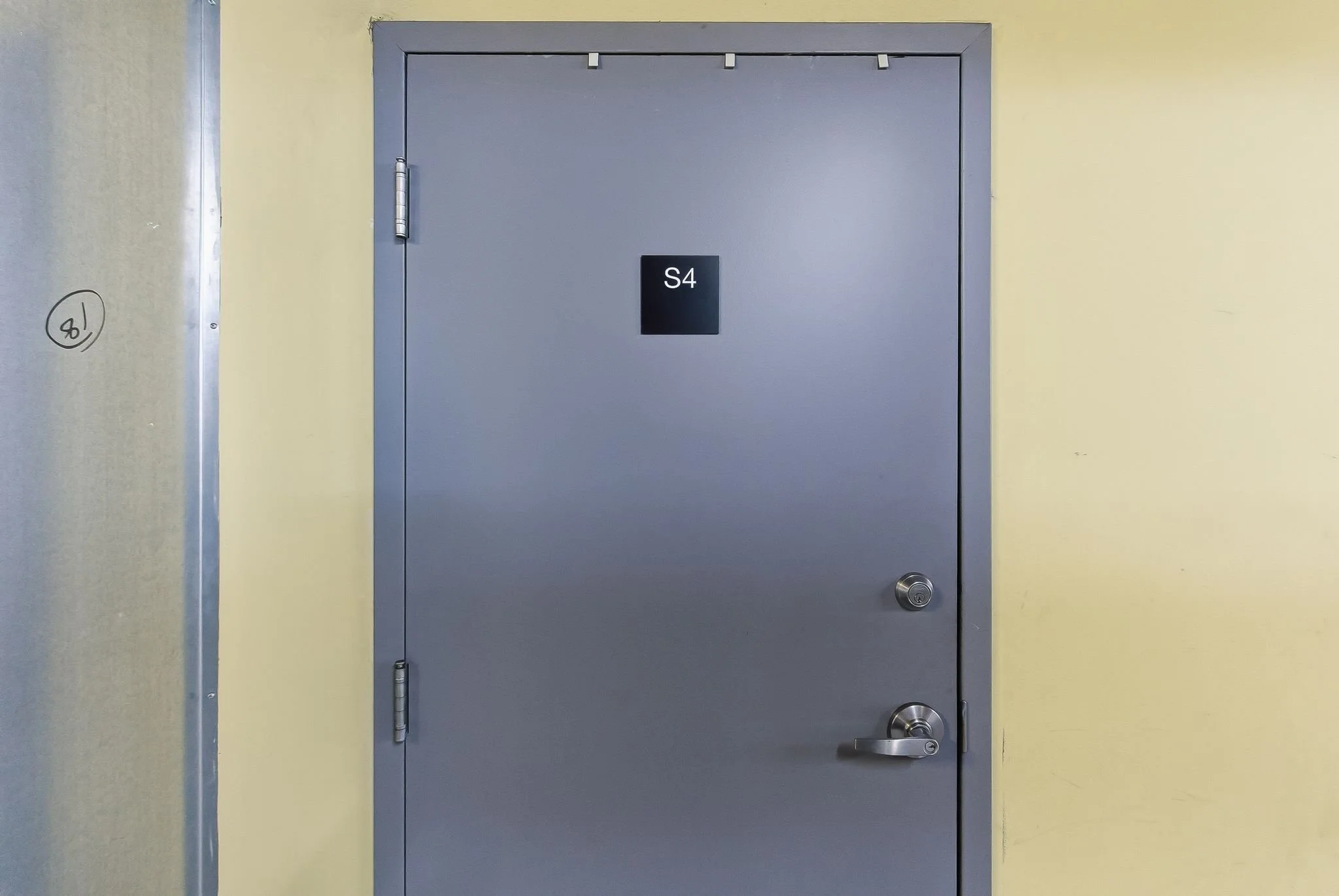 Gray door with a black sign labeled 'S4' located in the center. The door has a silver handle and a lock. The wall surrounding the door is painted light yellow.