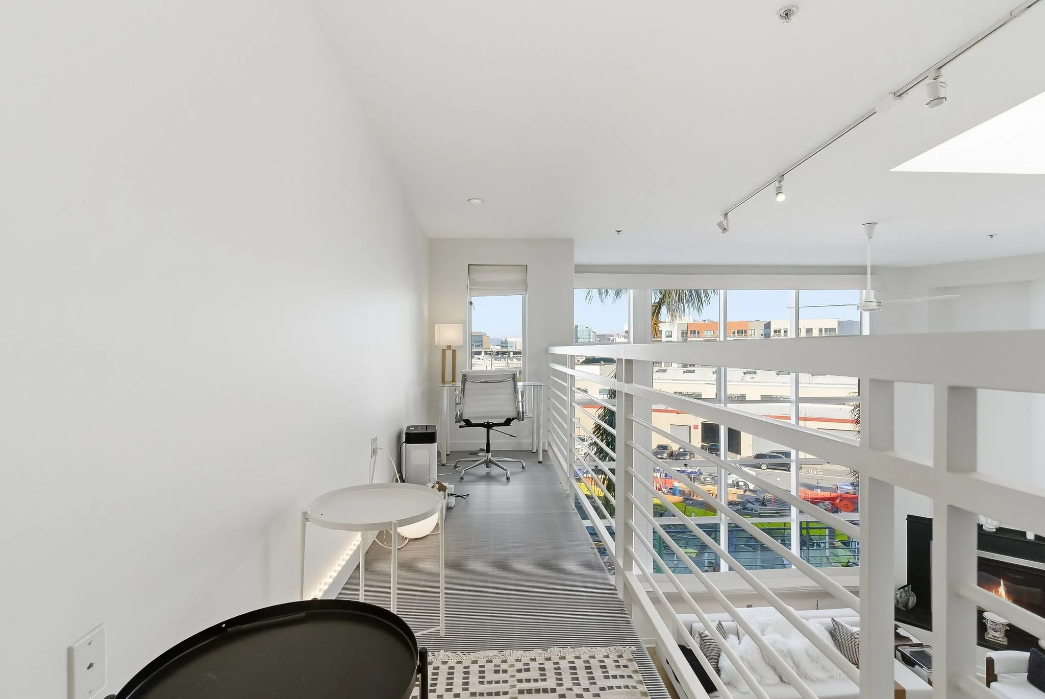 Interior view of a modern, bright space with a white railing, large windows overlooking a cityscape, a desk with an office chair, a lamp, and various small furniture pieces.