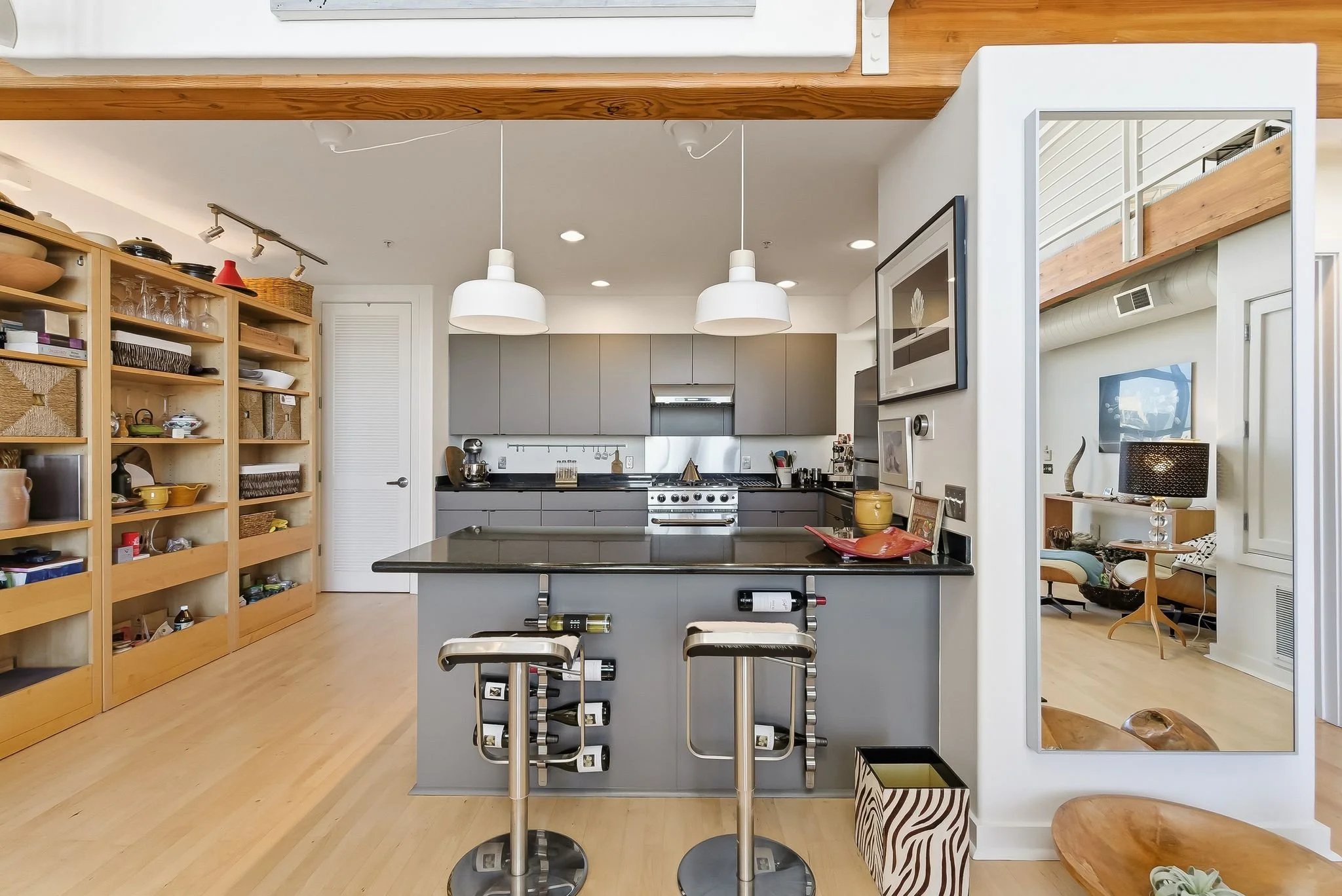 Modern kitchen with gray cabinets, black countertop island, open wooden shelves with dishes, two white pendant lights, and a view into a living room with a large wall mirror.
