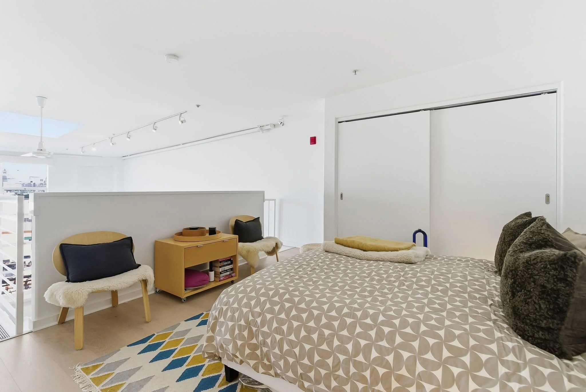 A modern, minimalist bedroom with a bed, cushions, small wooden furniture, and decorative rugs, in a bright and airy space with white walls and ceiling.