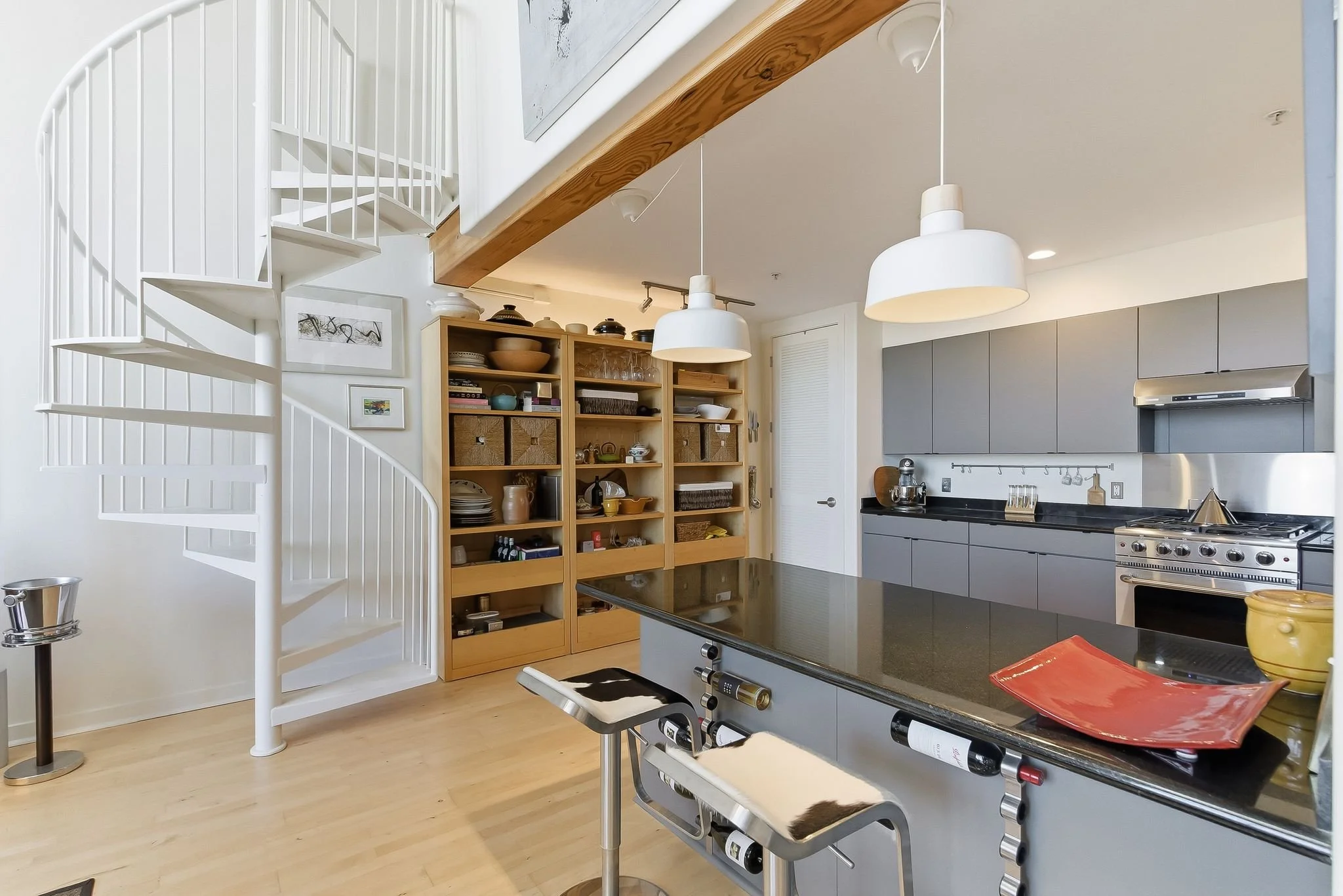 Modern kitchen with gray cabinets, black countertops, a white spiral staircase, and a wooden shelving unit filled with dishes and decor. Two white pendant lights hang over a kitchen island.