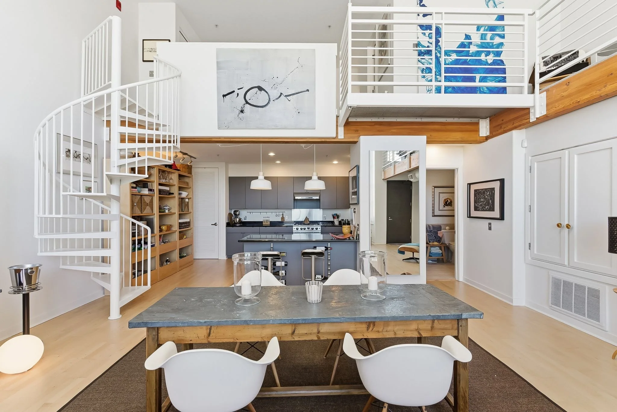 Open-concept living space with a dining table, modern kitchen, and loft area. The dining table has three candles and is surrounded by white chairs. The kitchen features gray cabinets and a small island with two stools. The loft has a white railing, w