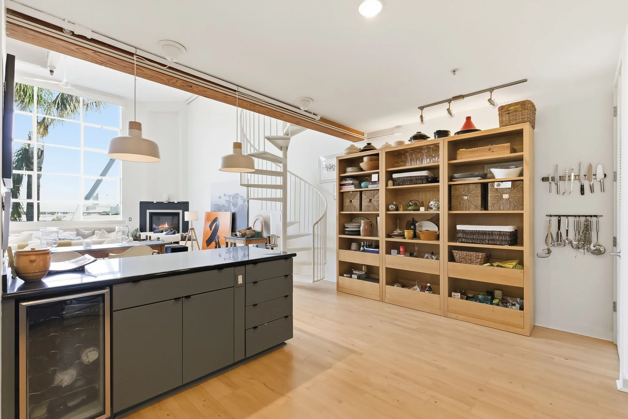 Open-concept kitchen and living room with large window, wooden shelving unit filled with dishes and kitchen items, and spiral staircase leading to upper level.